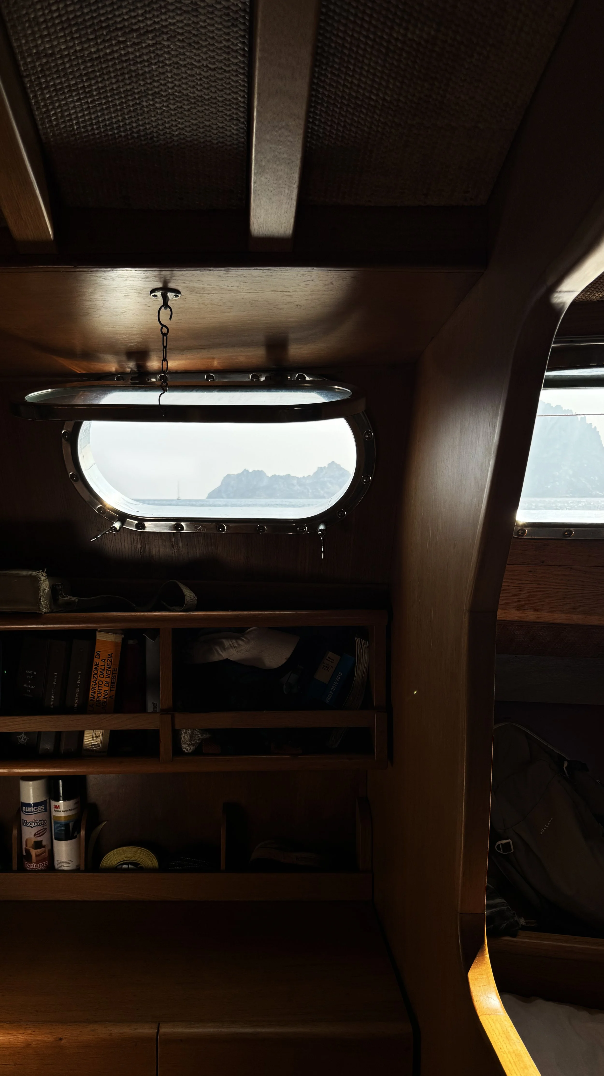 View of an island seen through an oval-shaped porthole window on a wooden boat cabin, with a bookshelf below the window containing books, tools, and supplies.
