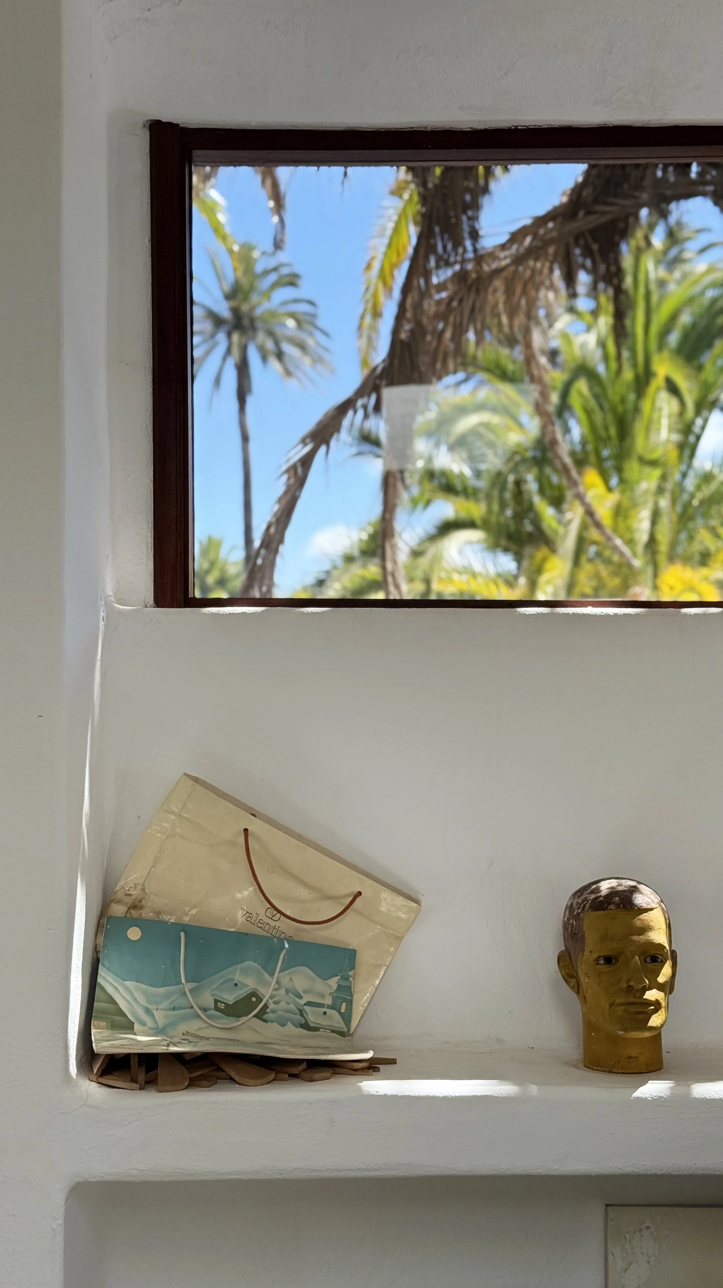 A window showing palm trees and a blue sky outside, with a paper bag and a decorative head sculpture on a white ledge inside.