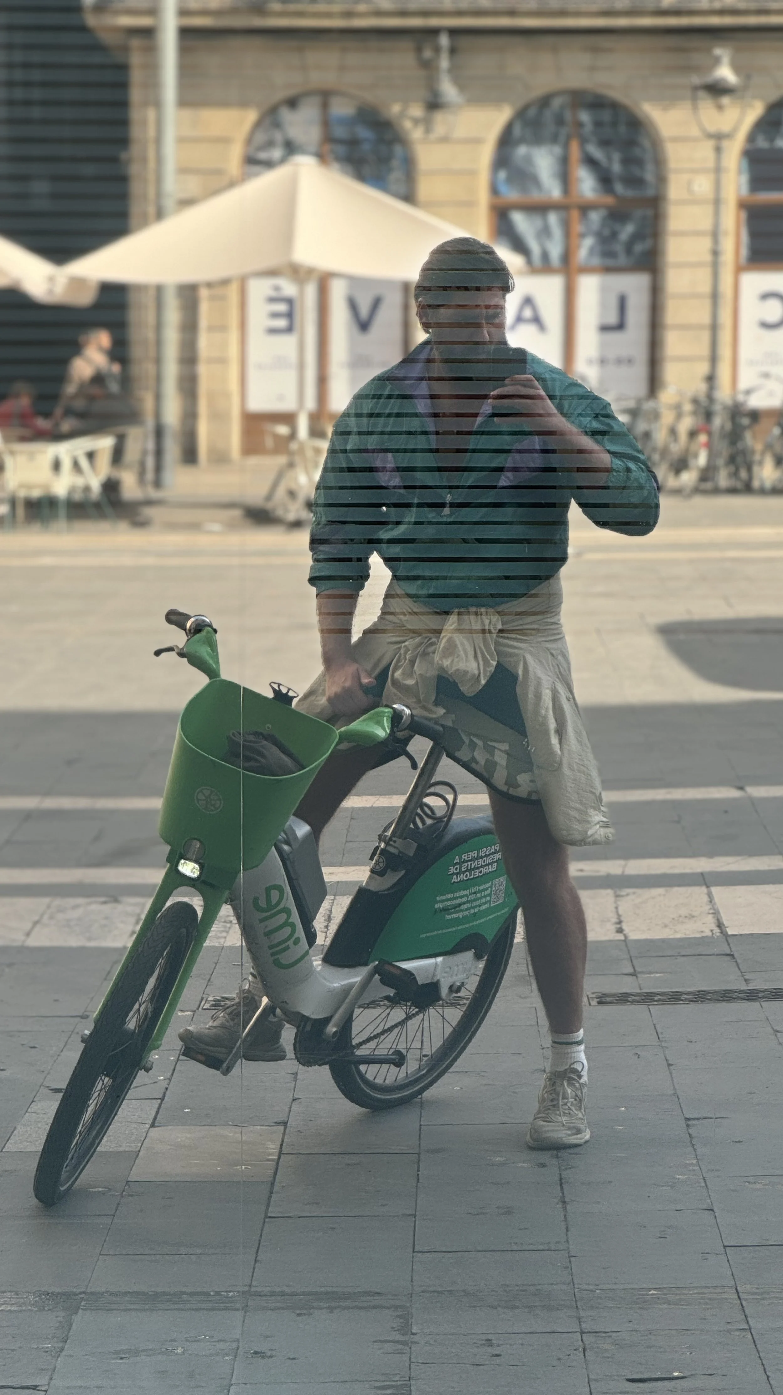 Benjamin Liatoud designs bespoke luxury architecture worldwide. Reflection of a man standing with a bike in front of a glass window on a city street.