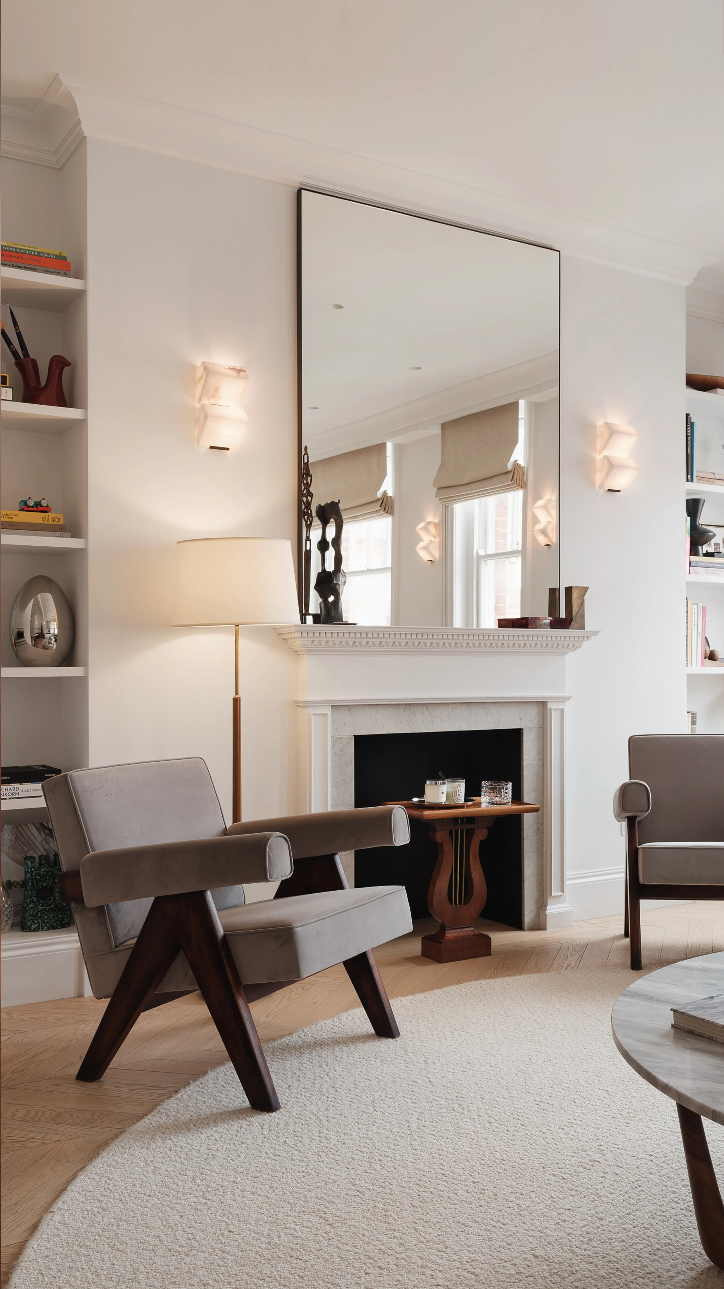 Living room with beige armchair, floor lamp, fireplace with decorative objects, large wall mirror, built-in bookshelves, and windows with beige Roman shades.