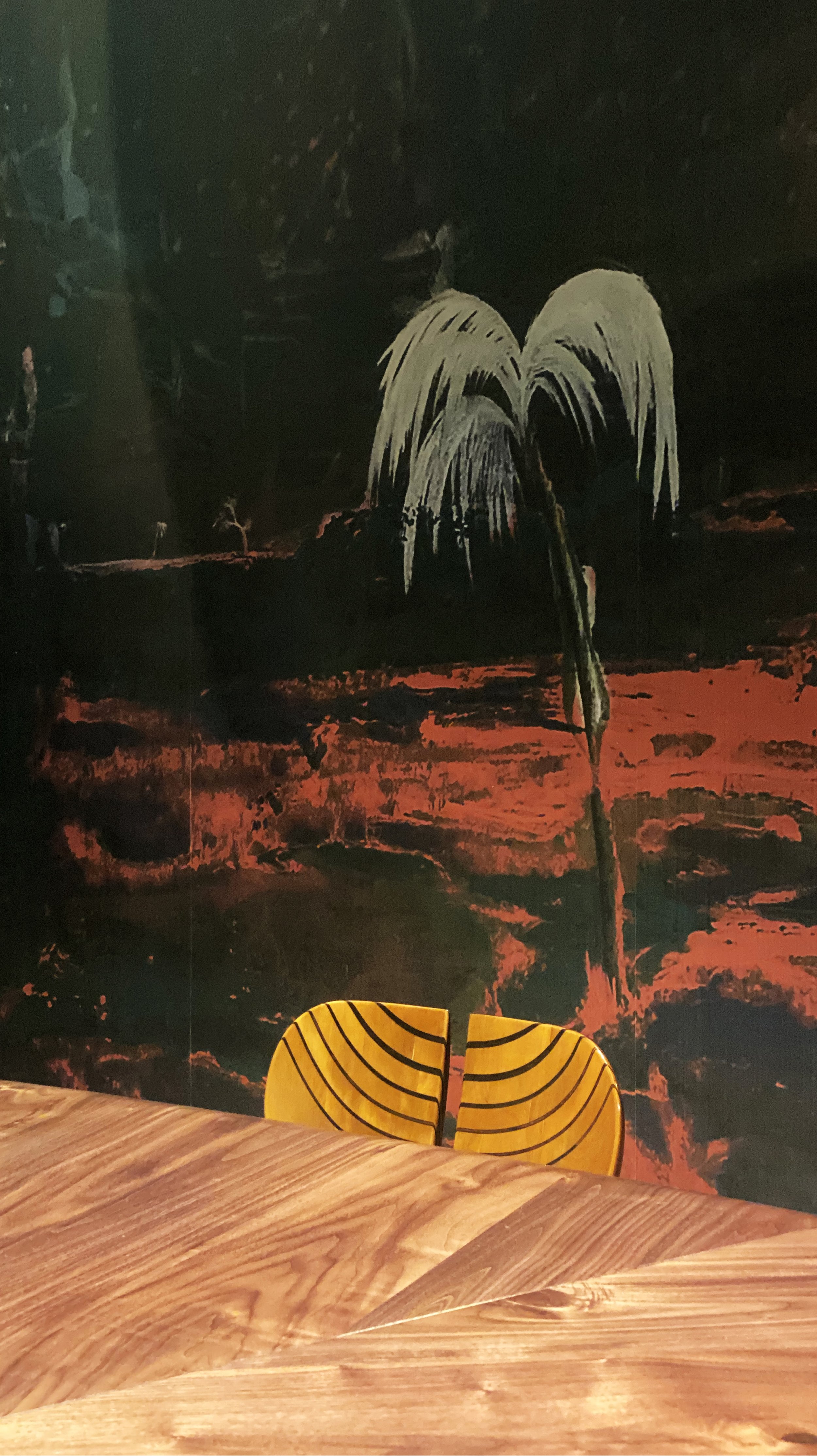 A yellow chair with black stripes placed behind a wooden table, set against a dark wall depicting a painted landscape with a tree and a fiery red ground.
