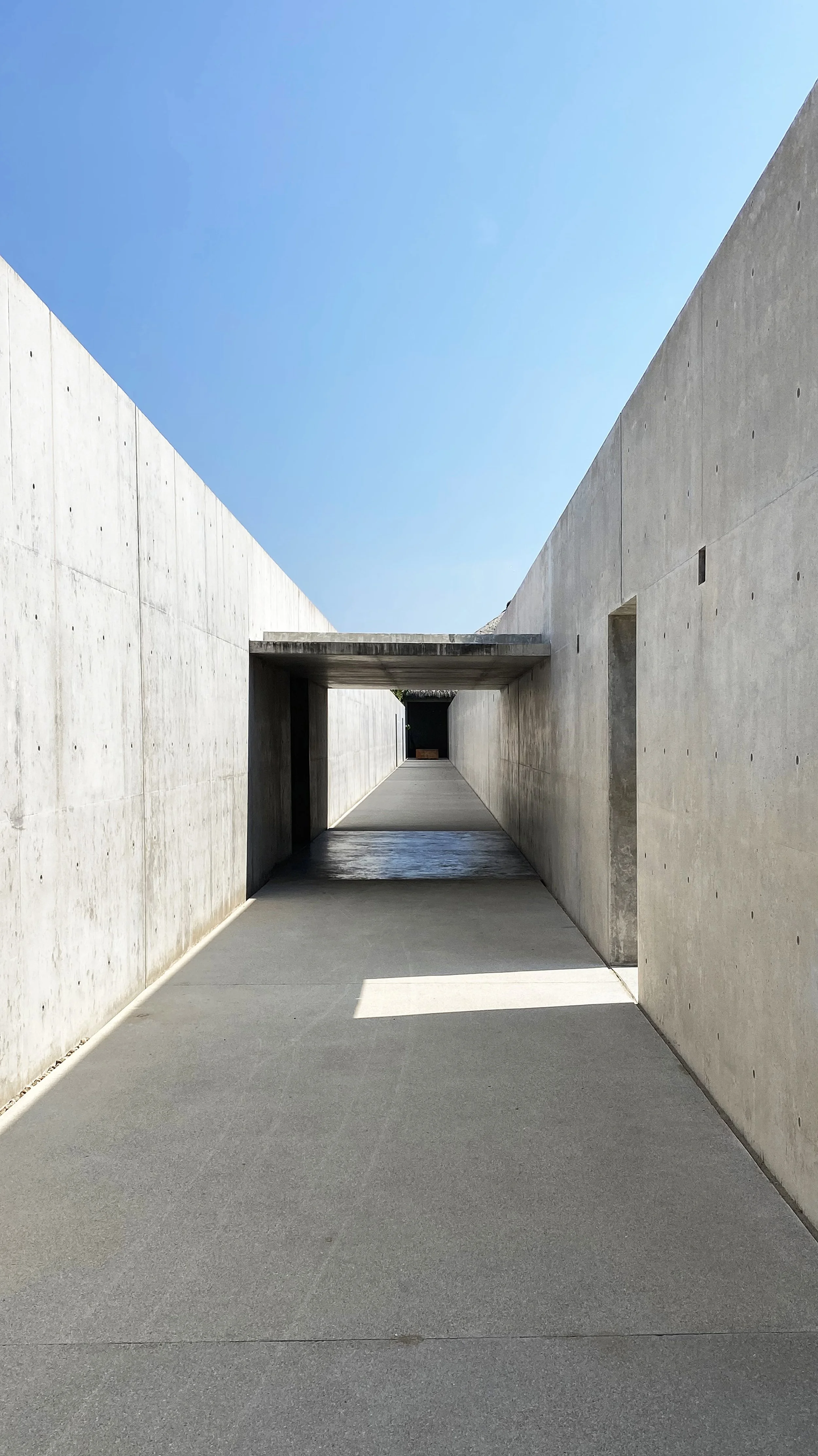 Photo of a modern concrete walkway flanked by tall concrete walls, with a rectangular tunnel overhead, leading into a space with a blue sky.