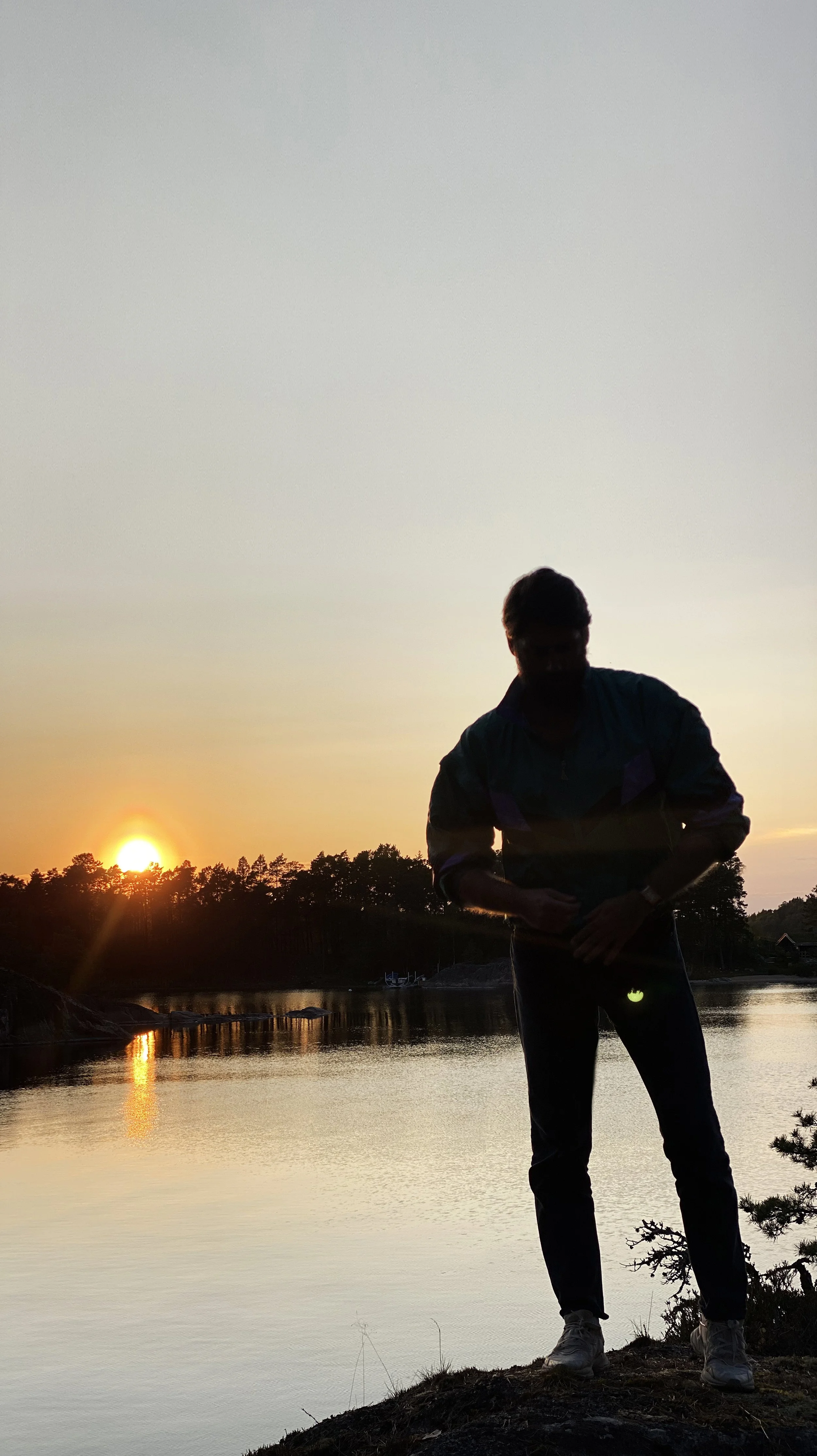 A silhouette of a person standing on a rock near water during sunset, with trees in the background.