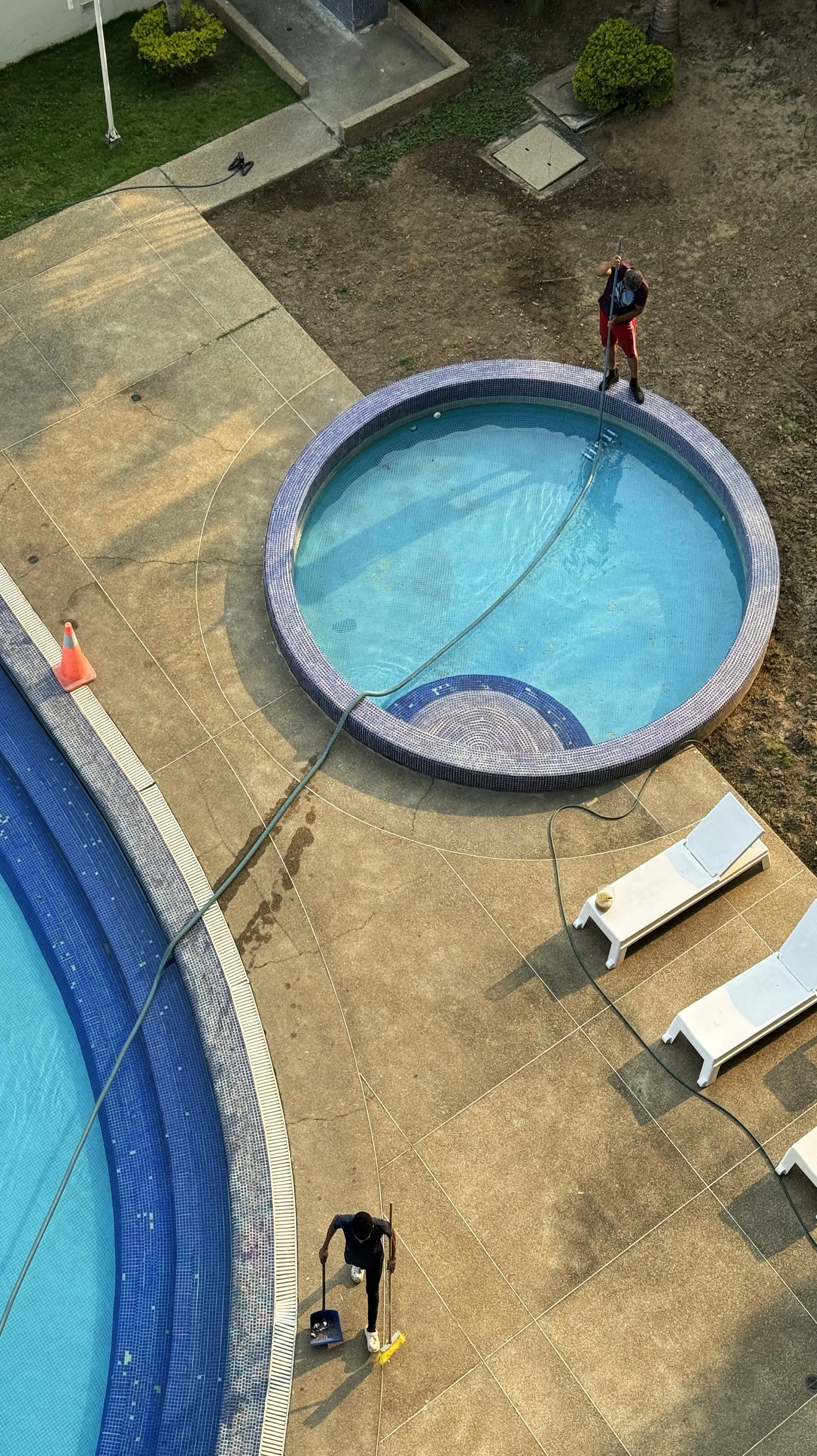 Two individuals are cleaning and maintaining a small round pool and a larger pool on a concrete patio. One person is using a long pole in the round pool, while the other has a mop and is near the larger pool.