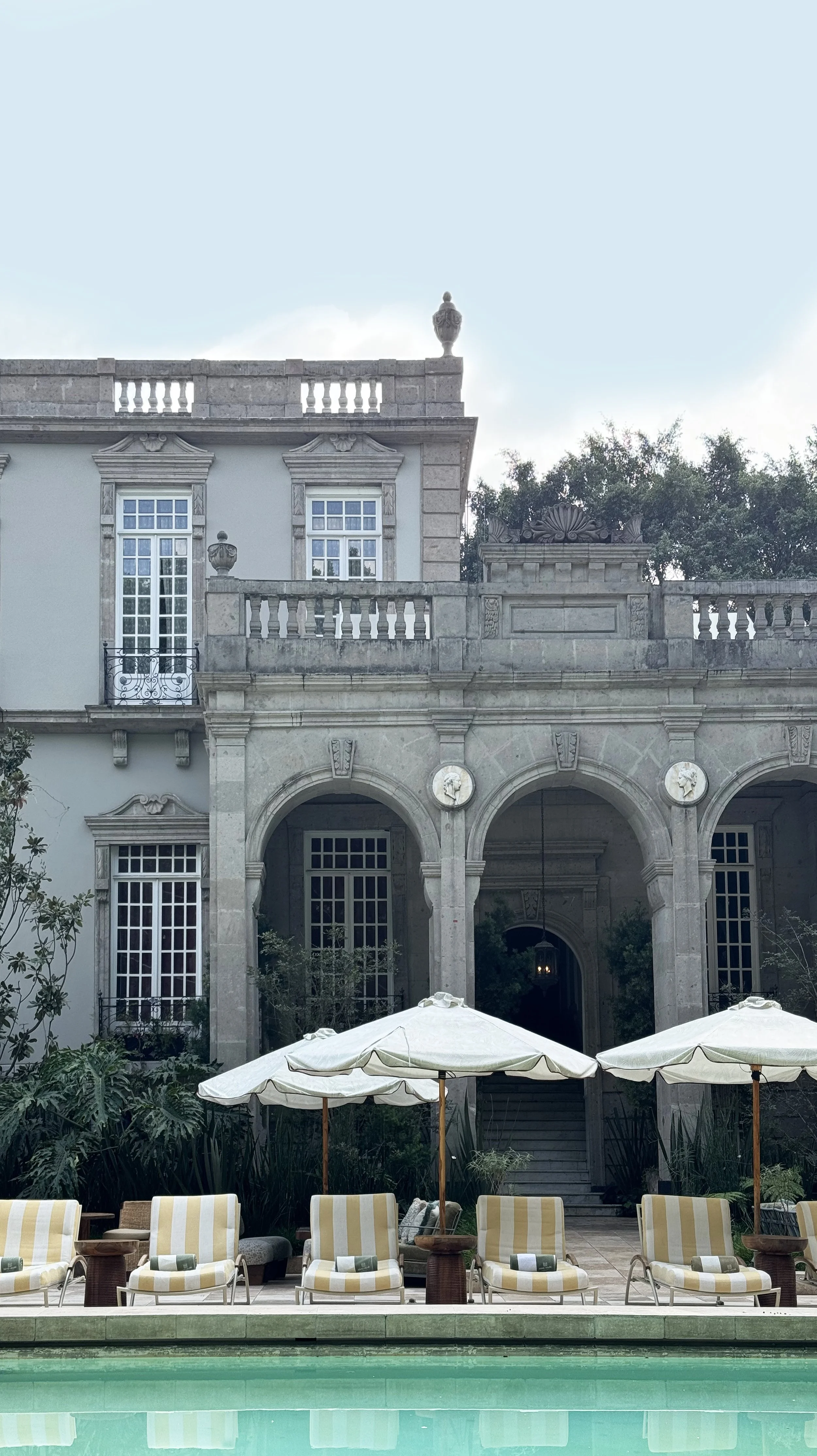 A luxurious mansion with classical architecture, stone arches, and large windows, with poolside lounge chairs and umbrellas in the foreground.