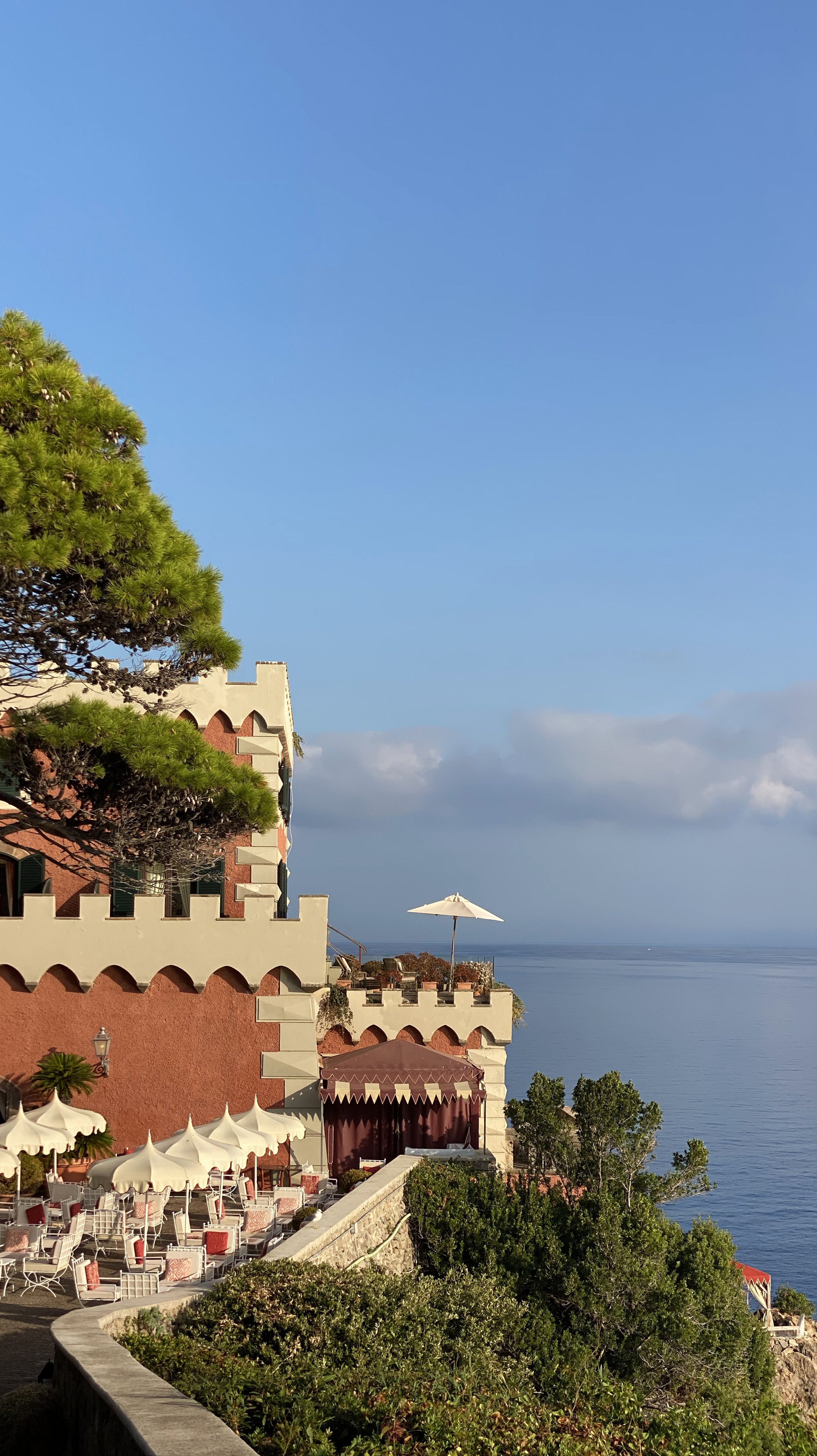 A coastal building with terraces, umbrellas, and outdoor seating overlooking the sea, with a blue sky and some clouds.