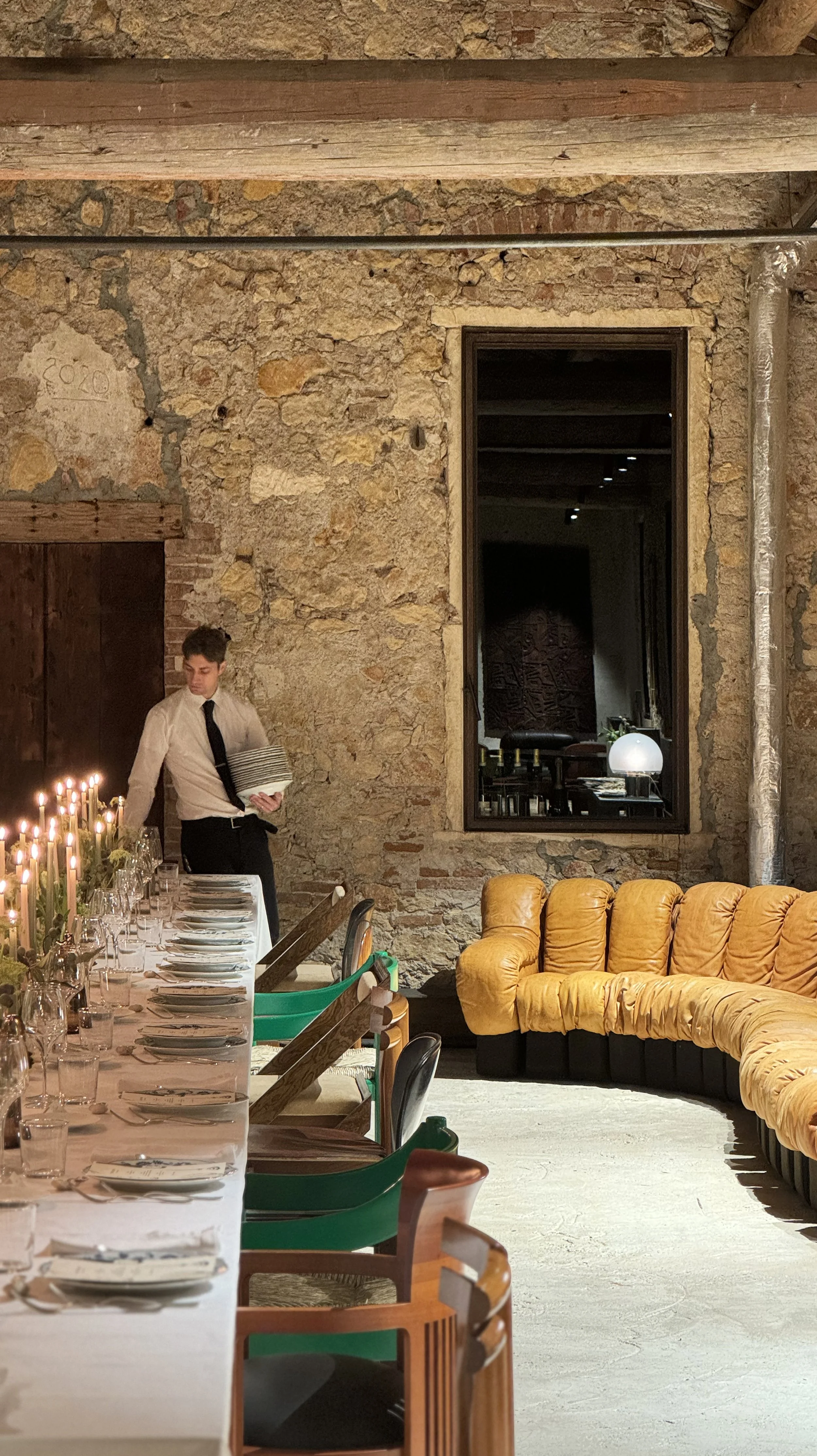 A man in a white shirt and black tie setting a long dining table with candles and glasses in a rustic restaurant with exposed brick walls, a large mirror, and a yellow tufted sofa.