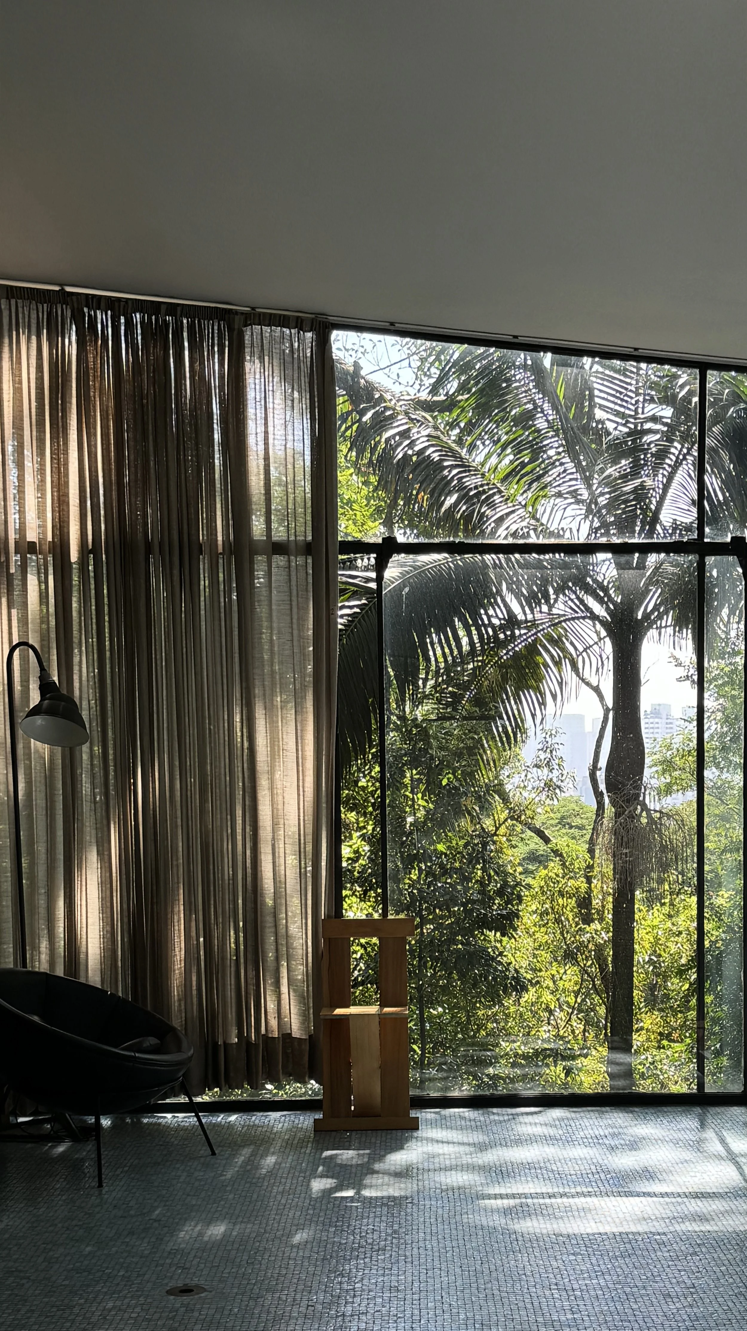 Indoor scene with large glass window revealing lush green trees outside, brown curtains, a black lounge chair, a wooden container, and a black lamp, sunlight casting shadows on the floor.