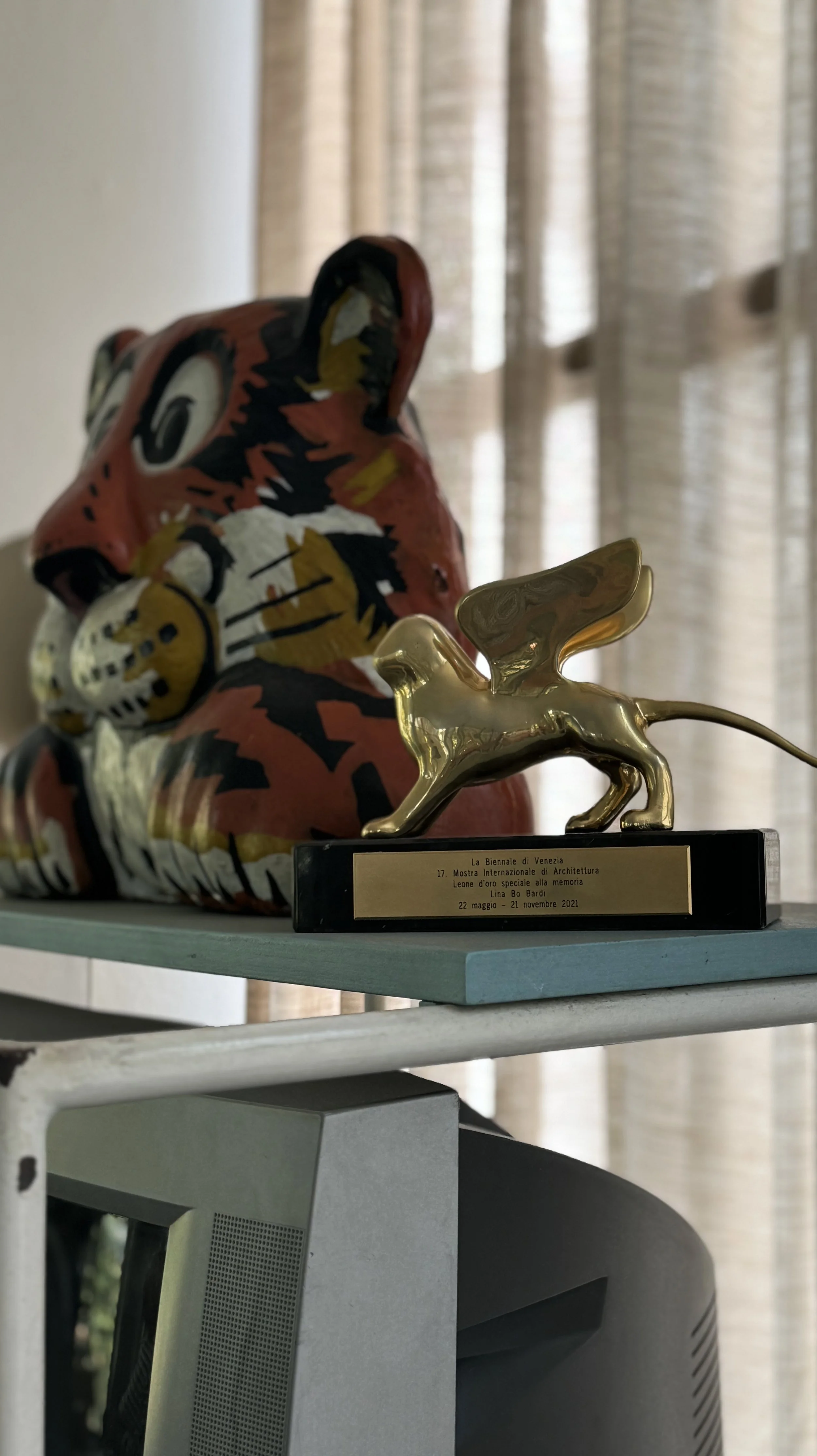 Decorative tiger sculpture with a white face and orange, black, and white striped body, and a golden lion figure with wings on a black base plate, placed on a table near a creased curtain.