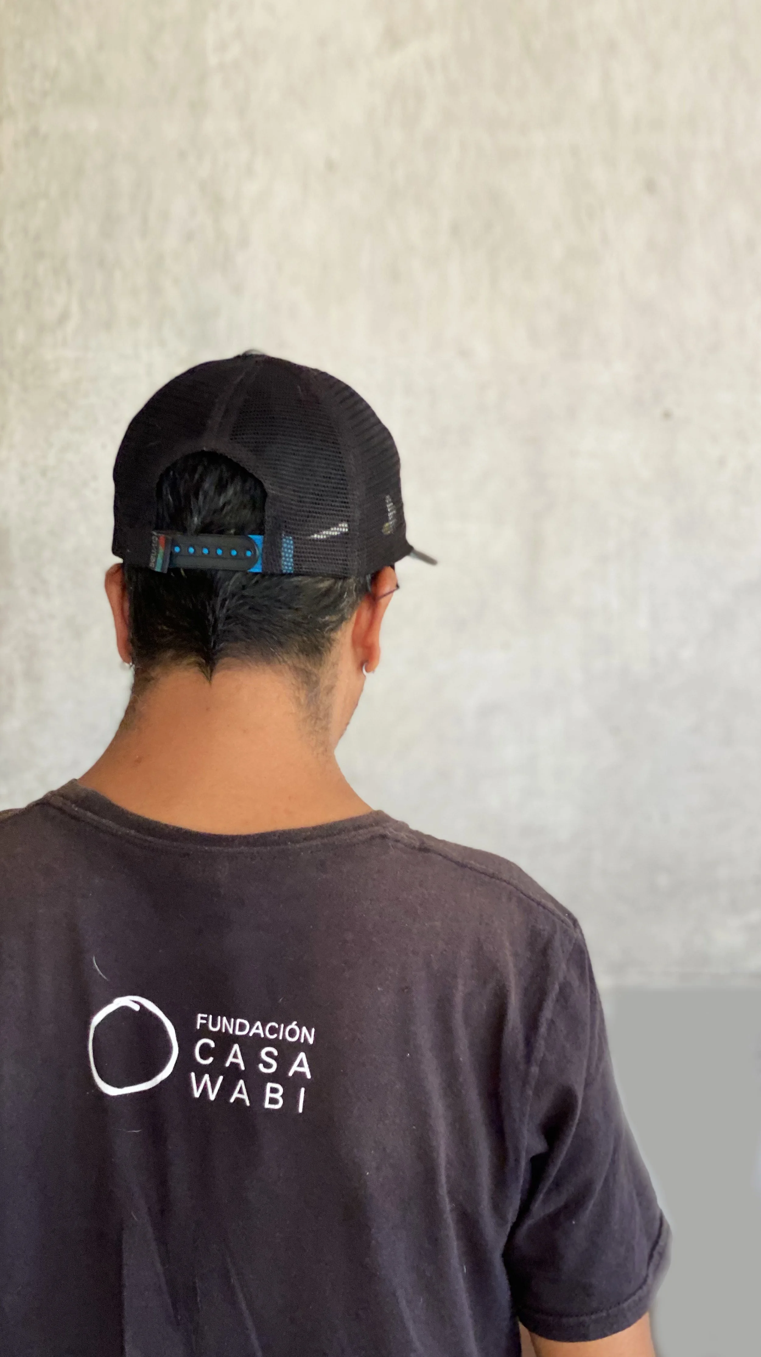Back of a person wearing a black baseball cap and a black T-shirt with white text. The cap has a blue strap, and the person's hair is visible underneath. The T-shirt has the logo and text 'Fundación Casa Wabi' on the back.