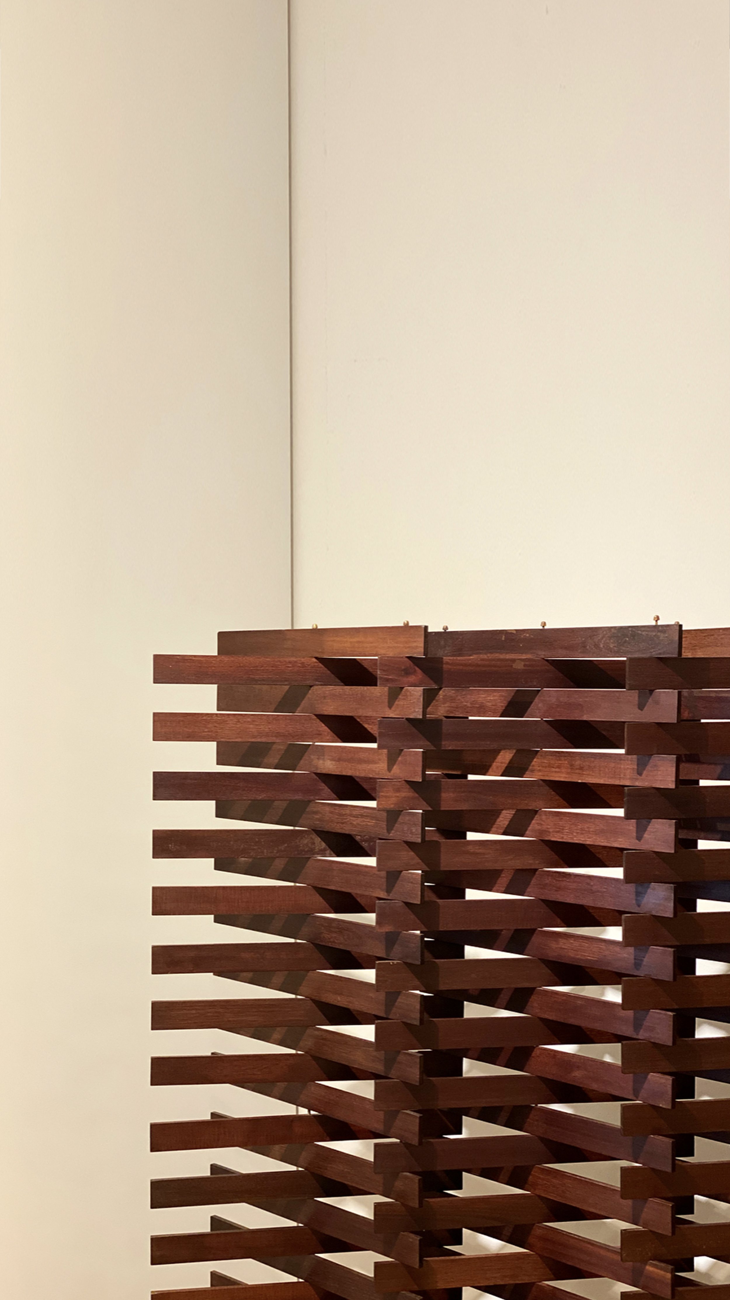 A tall, modern wooden sculpture made of multiple horizontal pieces arranged in an intricate, geometric pattern, placed against a plain, light-colored wall.