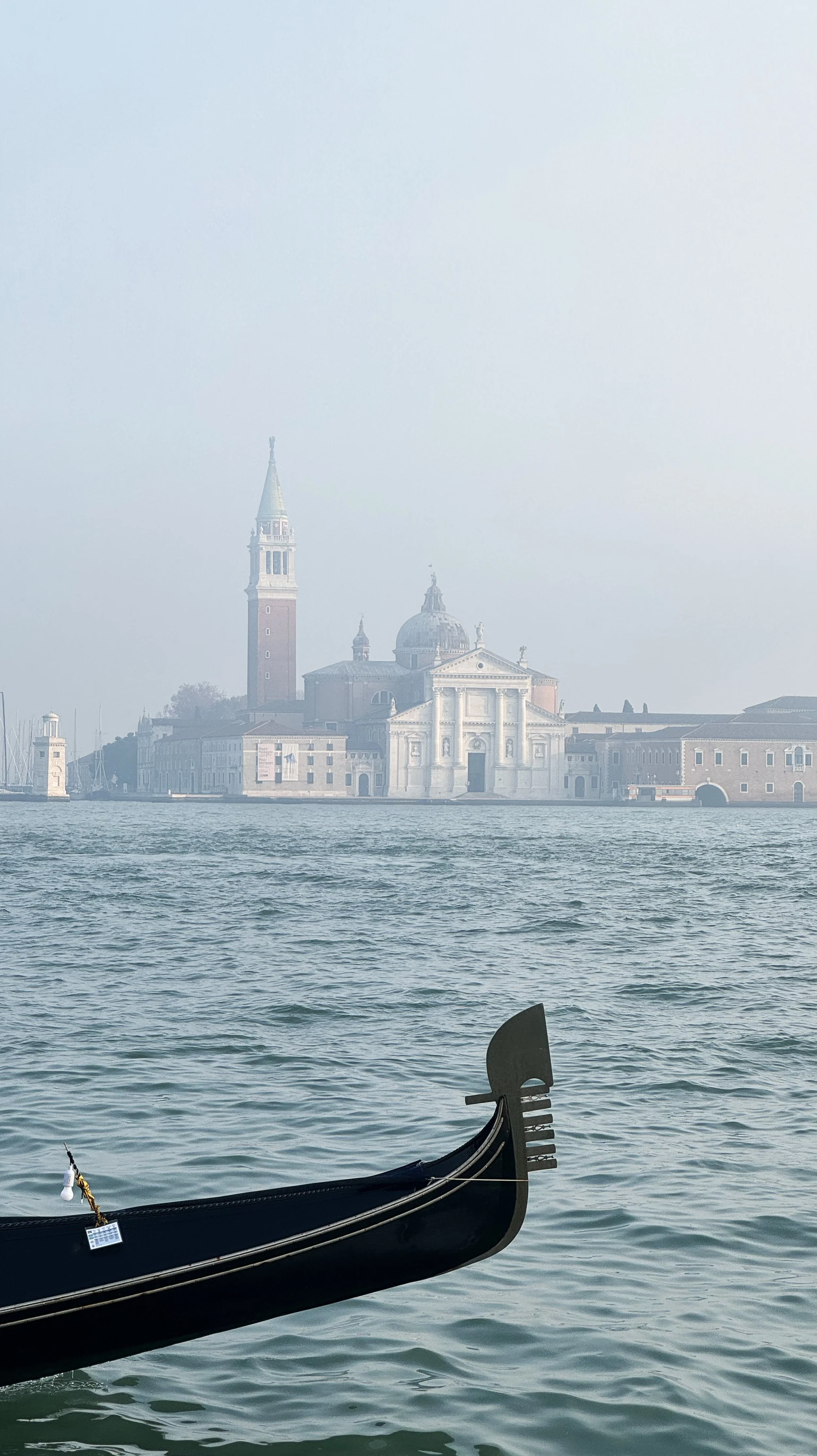 A gondola on the water with a skyline of historic buildings and a church with a tall bell tower in the background, shrouded in fog.