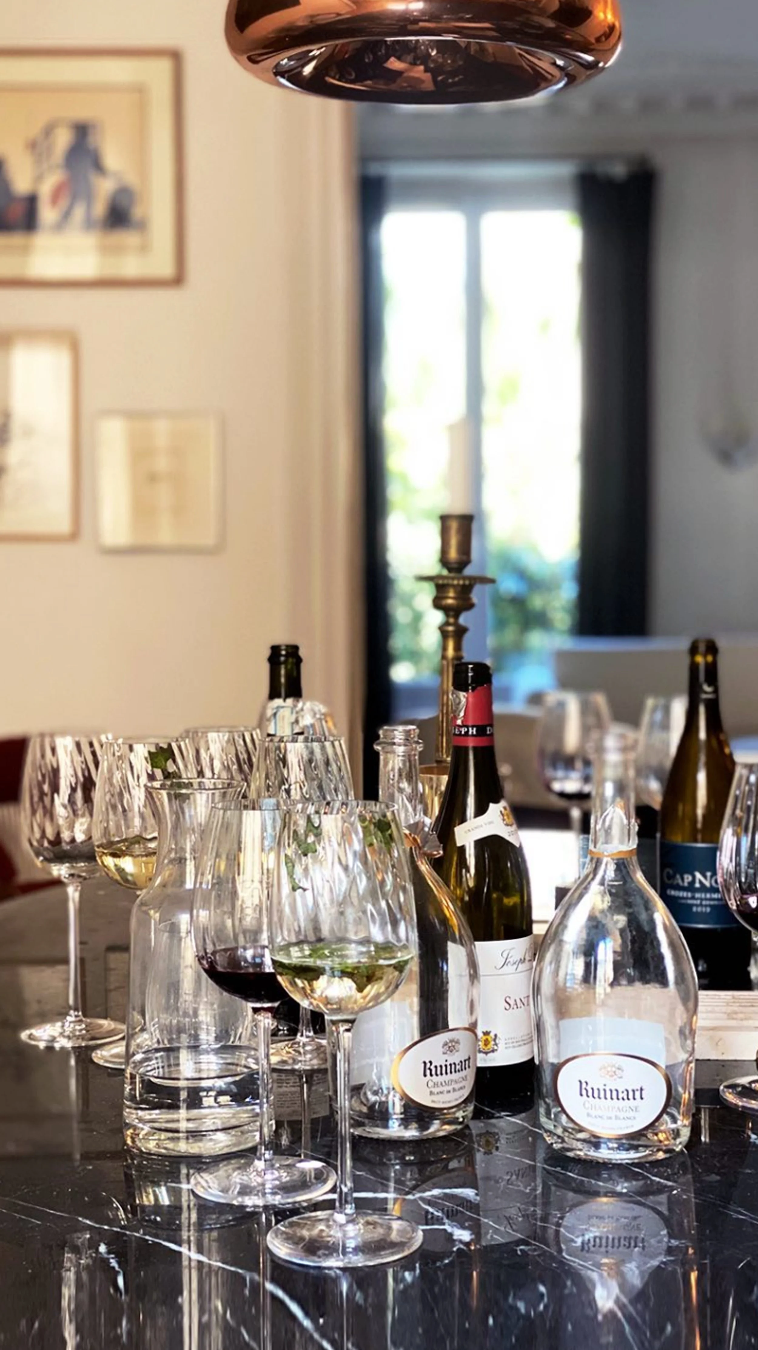 A dining table with multiple wine glasses, some filled with white and red wine, and several bottles of wine. A candlestick is also on the table, and a window with curtains is in the background, letting in natural light.
