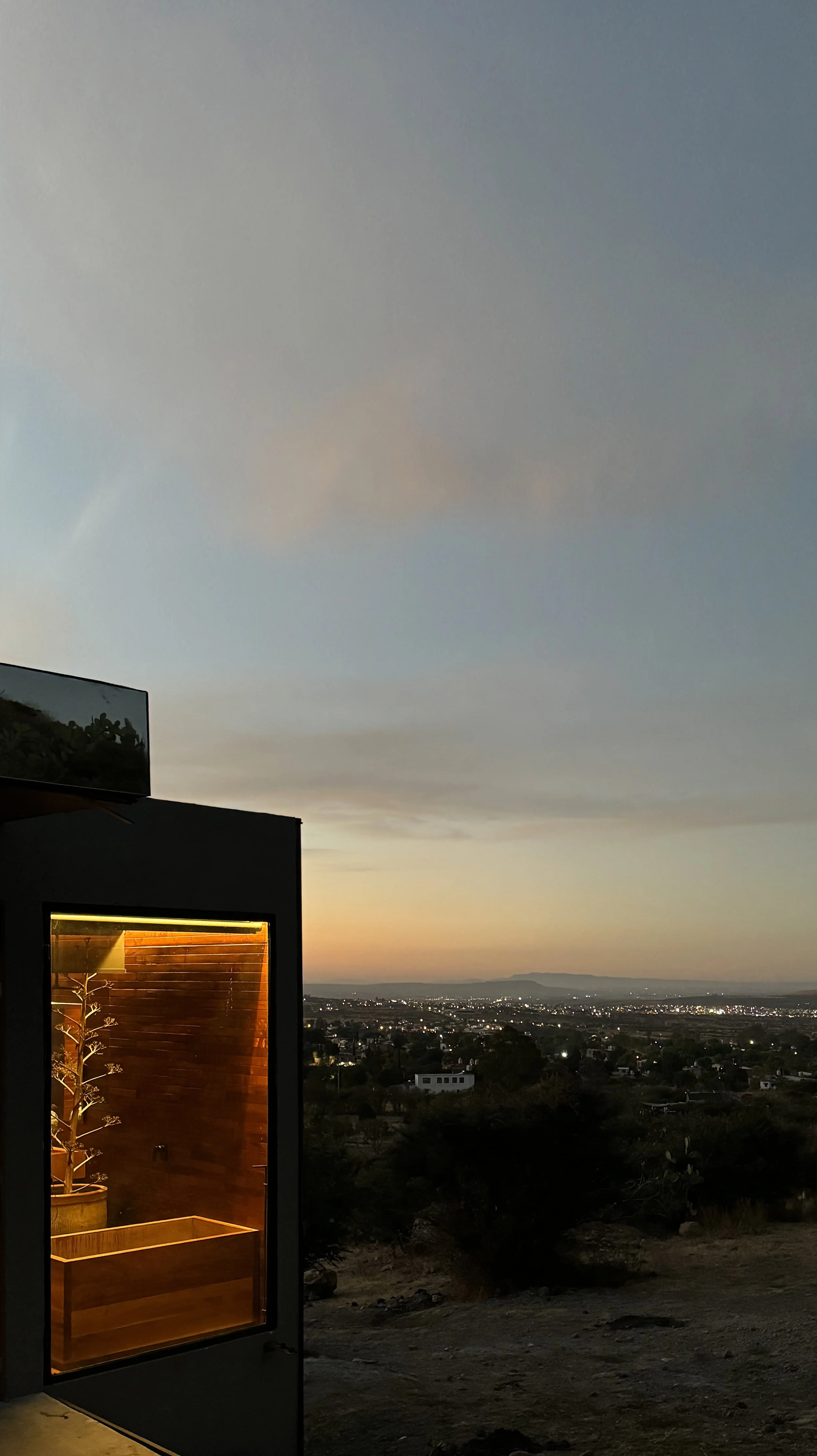 A sunset view with a cityscape in the distance, a dark building with a lit interior featuring a plant in a pot on a wooden surface, and a cloudy sky overhead.