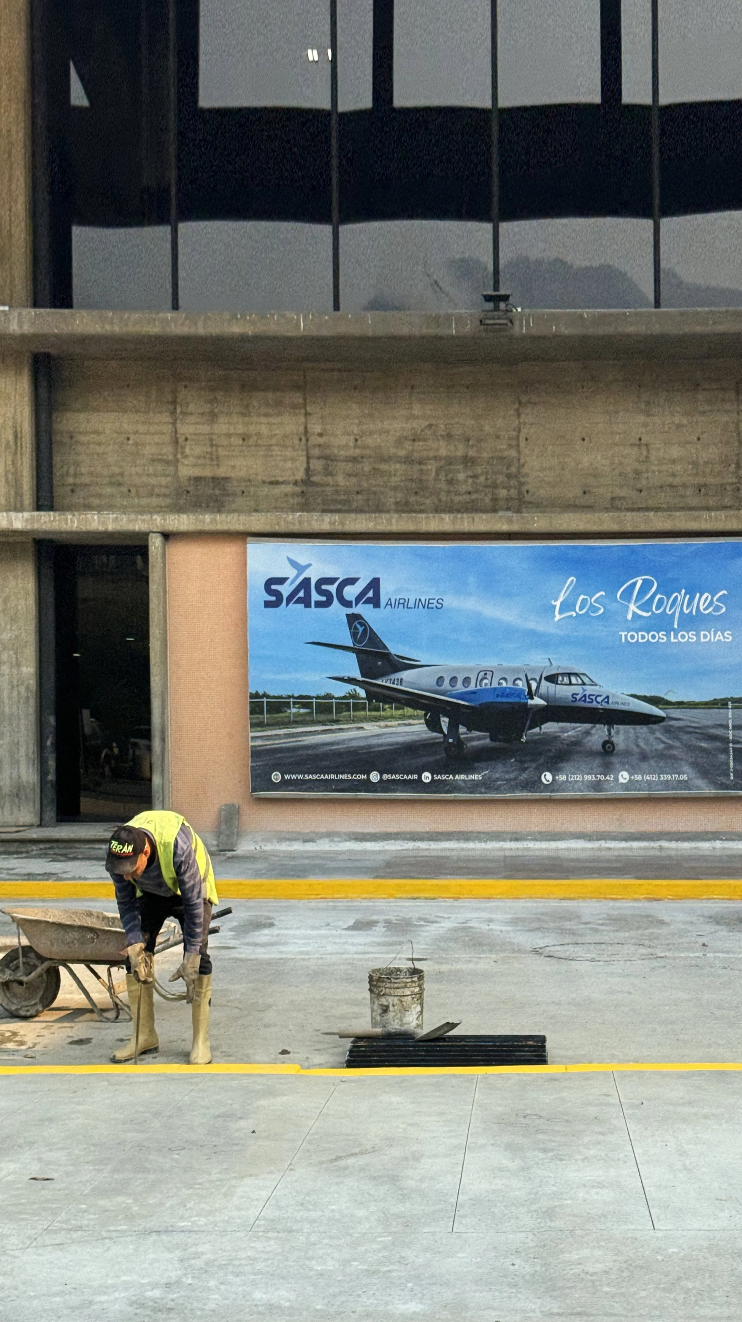 Construction worker laying concrete on a sidewalk in front of a SASCA Airlines billboard.