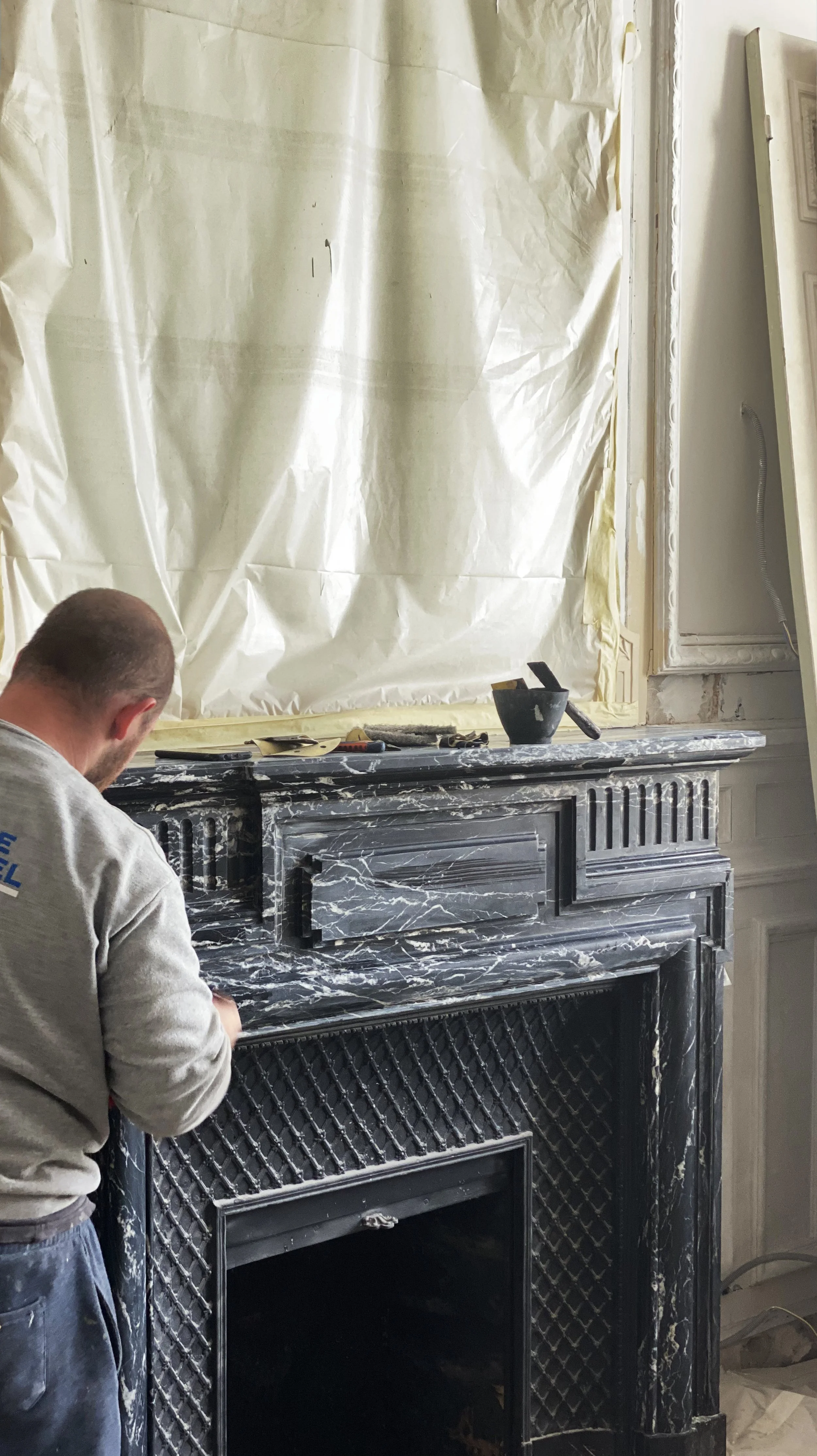 A man working on a black marble fireplace surround in a room under renovation, with plastic sheeting covering the wall behind him.