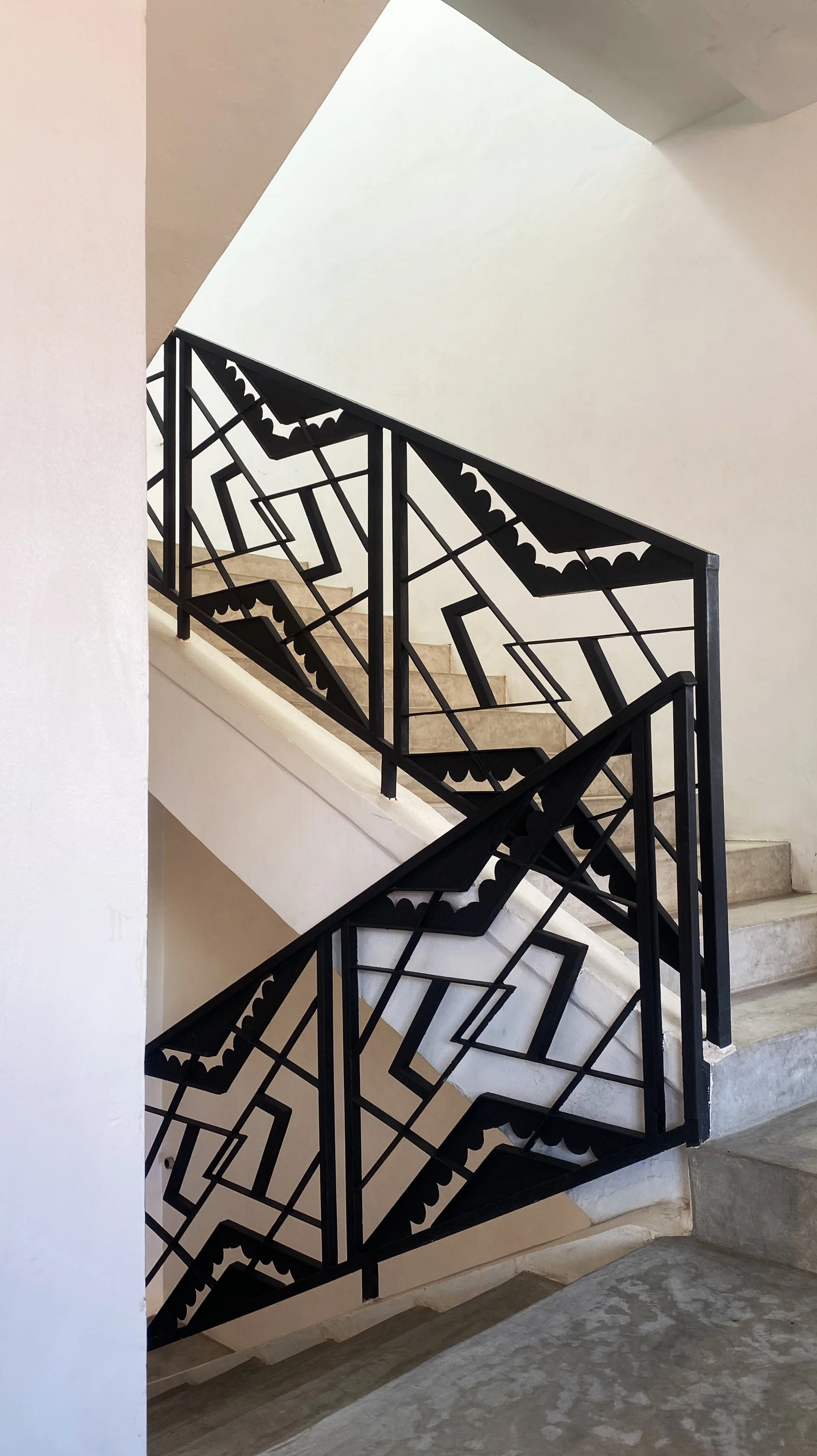 Decorative black metal staircase railing with geometric and bat-shaped patterns, adjacent to beige tiled stairs and white walls, in a modern building.