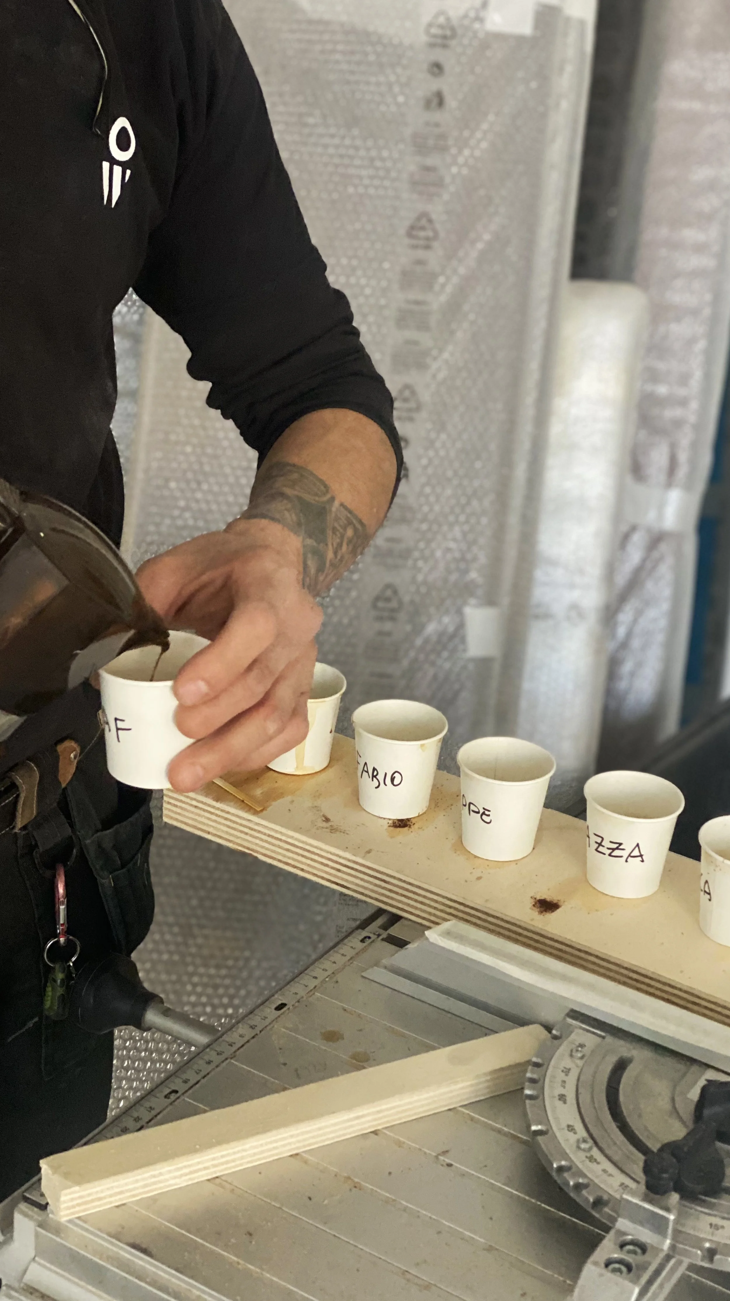 A person pouring coffee into paper cups labeled with names on a wooden board, on a worktable with a circular saw.