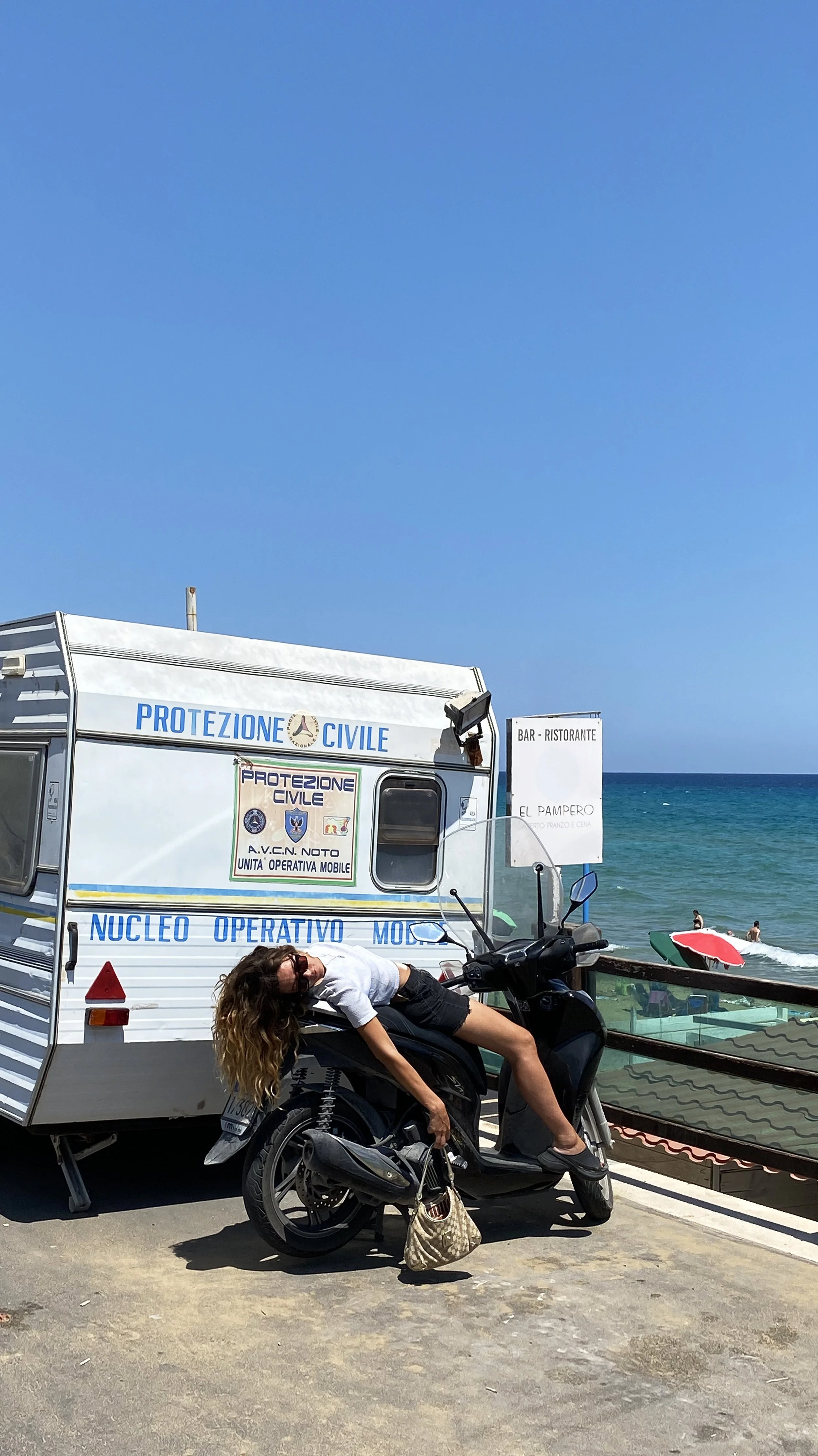 Woman with curly hair wearing sunglasses, white shirt, and black shorts, leaning on a black scooter by a blue and white mobile unit with blue text, near the ocean with beach umbrellas and people in the background.