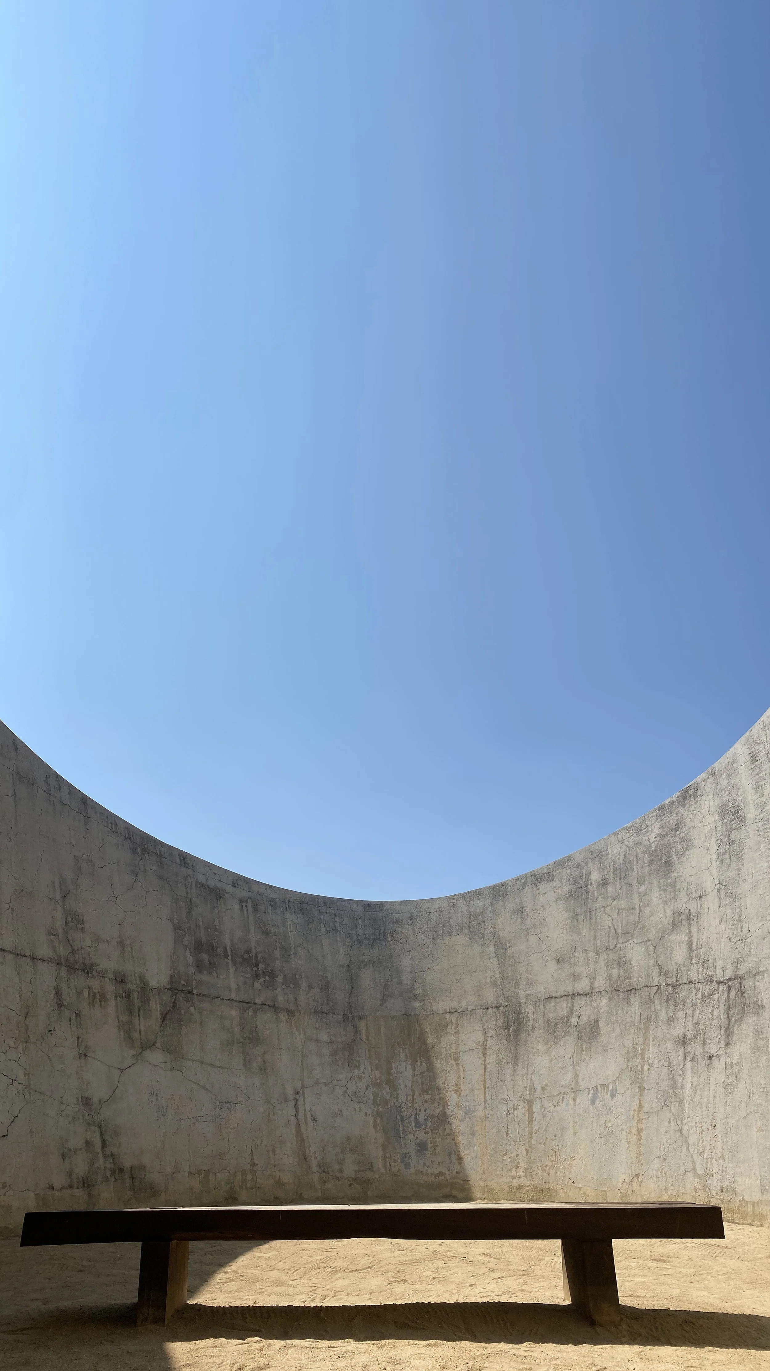 A low wooden bench set on sandy ground inside a concrete circular structure with an open top, revealing a clear blue sky.