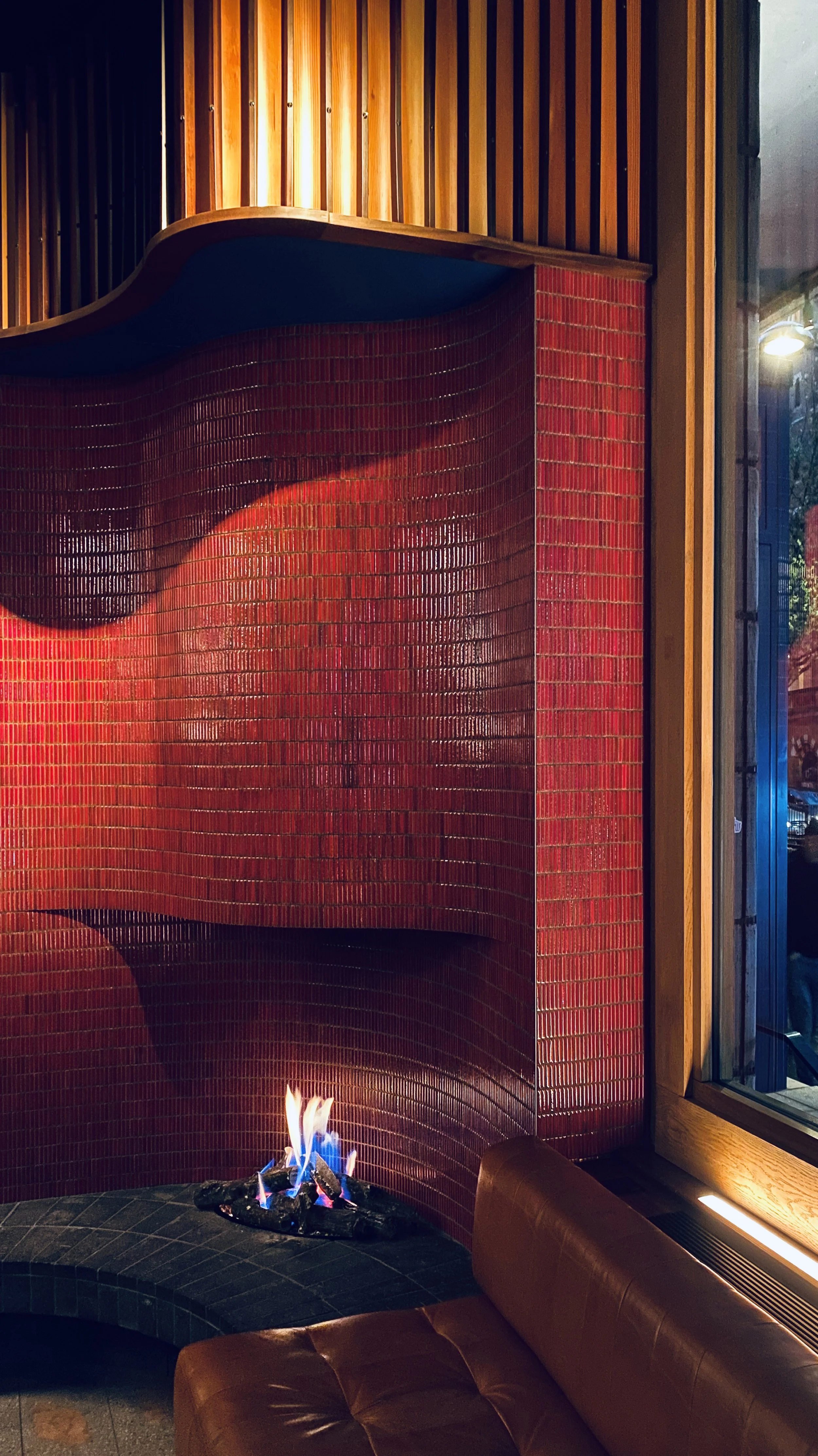 Interior of a modern lounge area with a small gas fireplace, red mosaic tile wall, wooden accents, large window showing outdoor scene, and a brown leather chair.