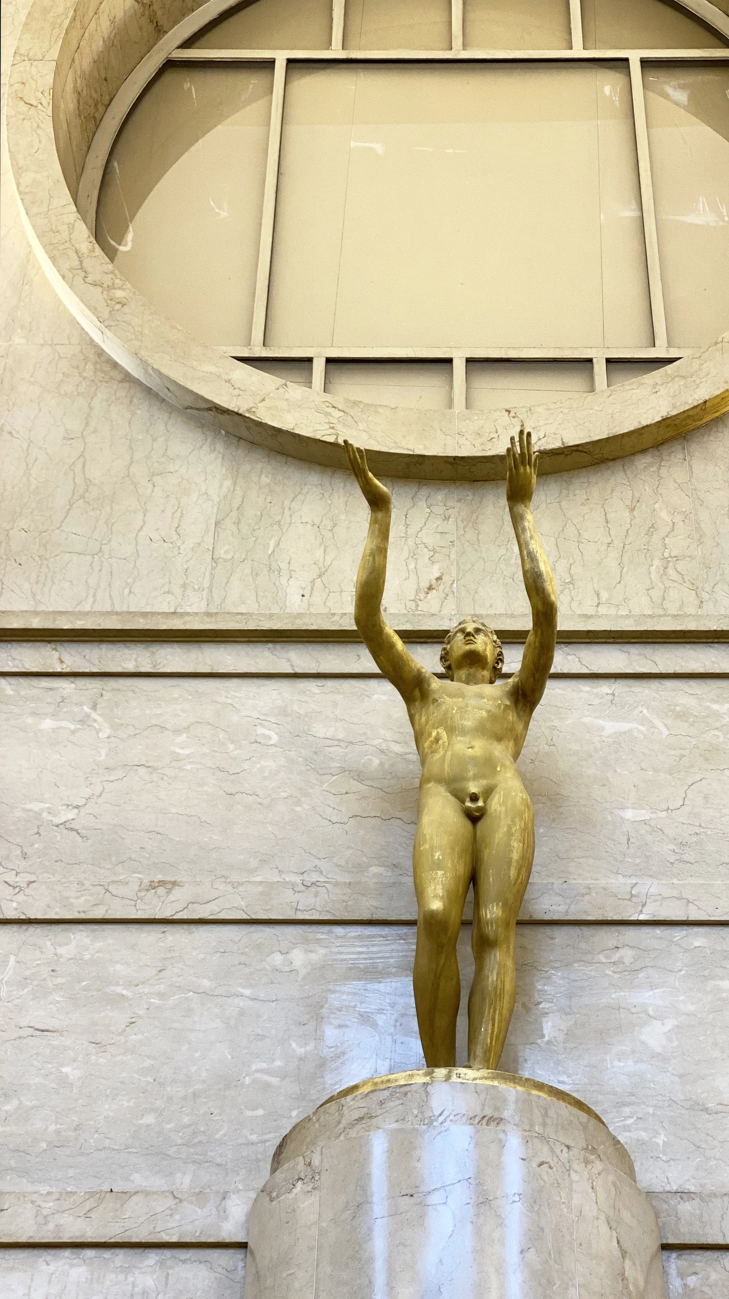 A golden statue of a nude child with arms raised above his head, standing on a marble pedestal inside a building with marble walls, with a large decorative window or opening above.