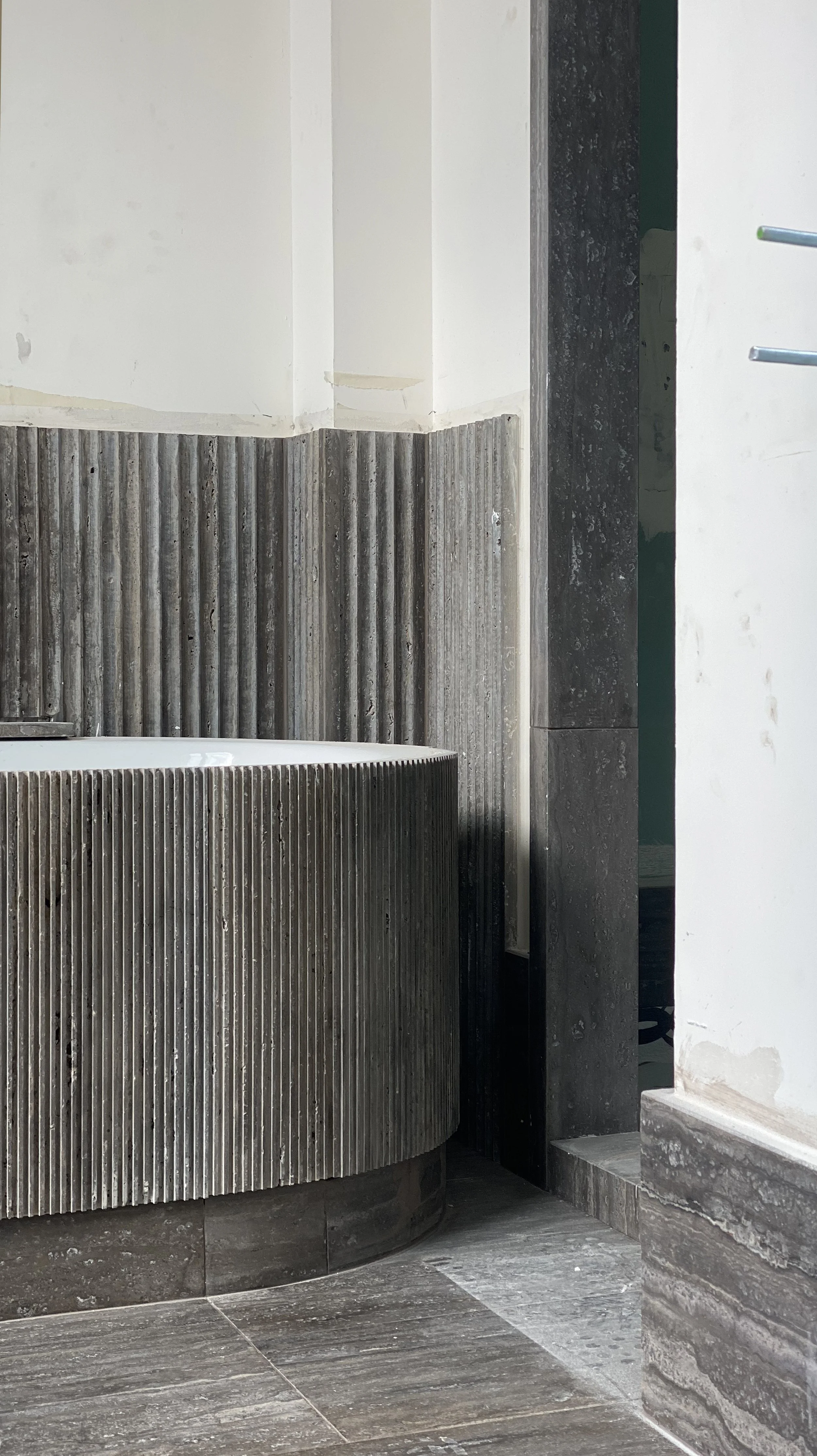 Part of a bathroom with a curved bathtub with ribbed vertical wood slats, dark tiles on the floor and wall, and a white wall with some visible patches.