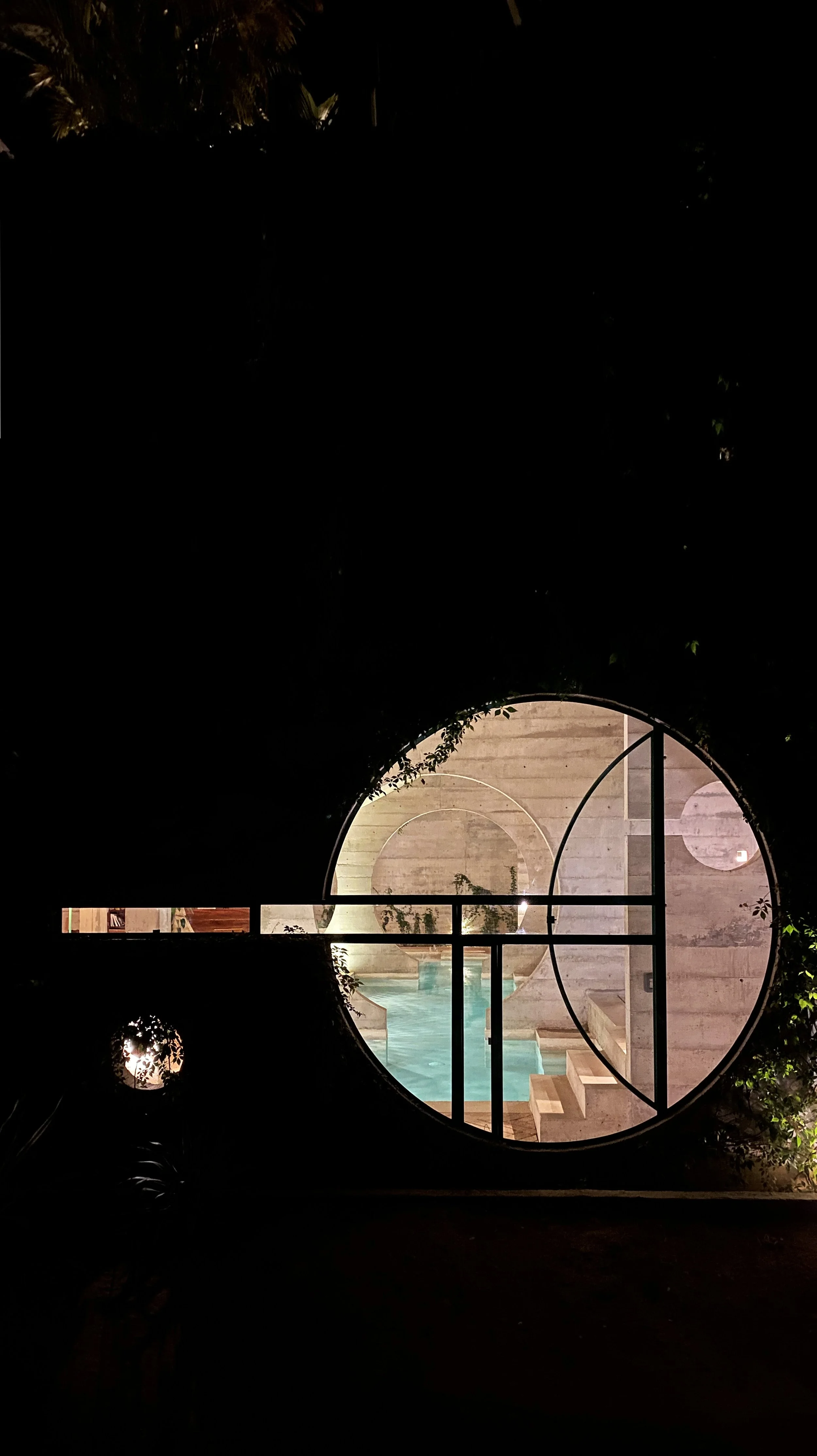 Night view of a swimming pool framed by a circular window with geometric design, surrounded by plants and dim lighting.