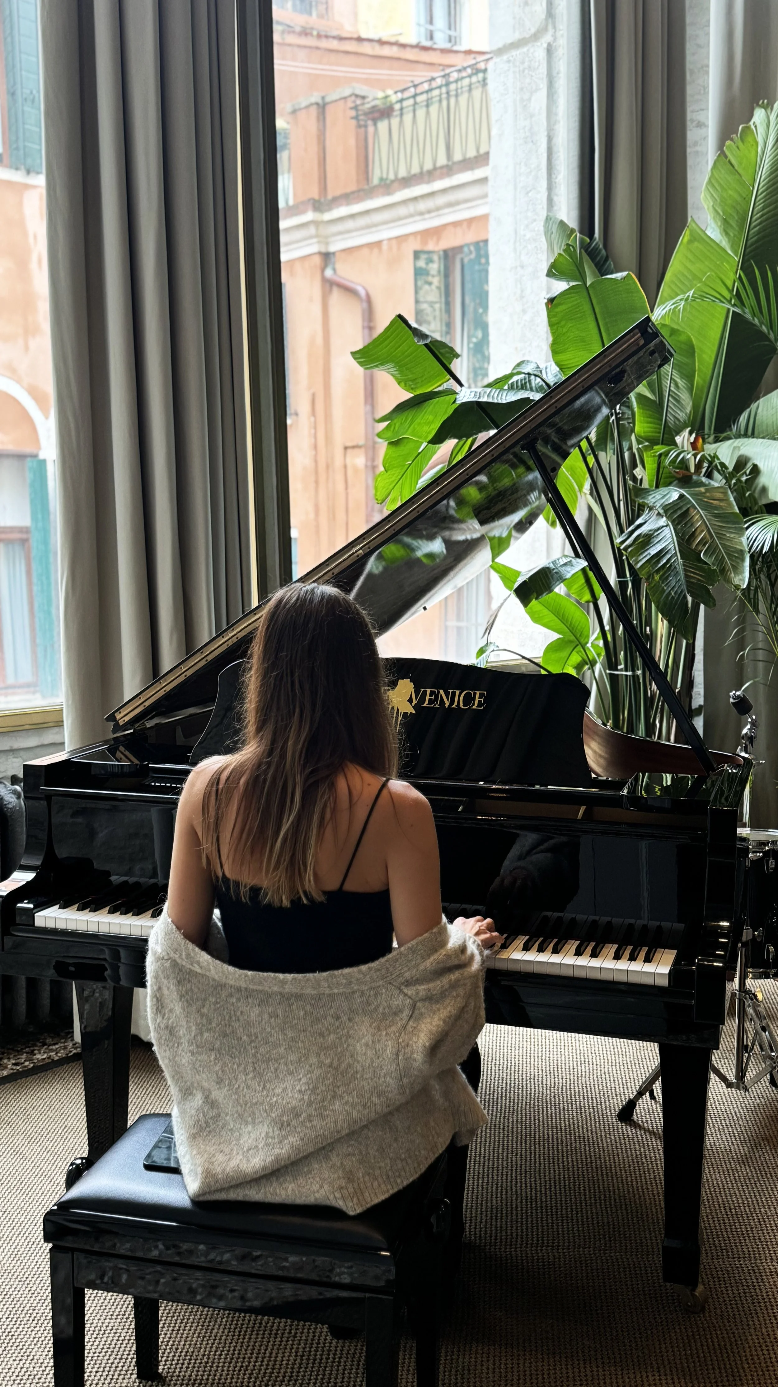 A woman with long brown hair seated at a black grand piano by a large window with green plants nearby, looking at the piano.