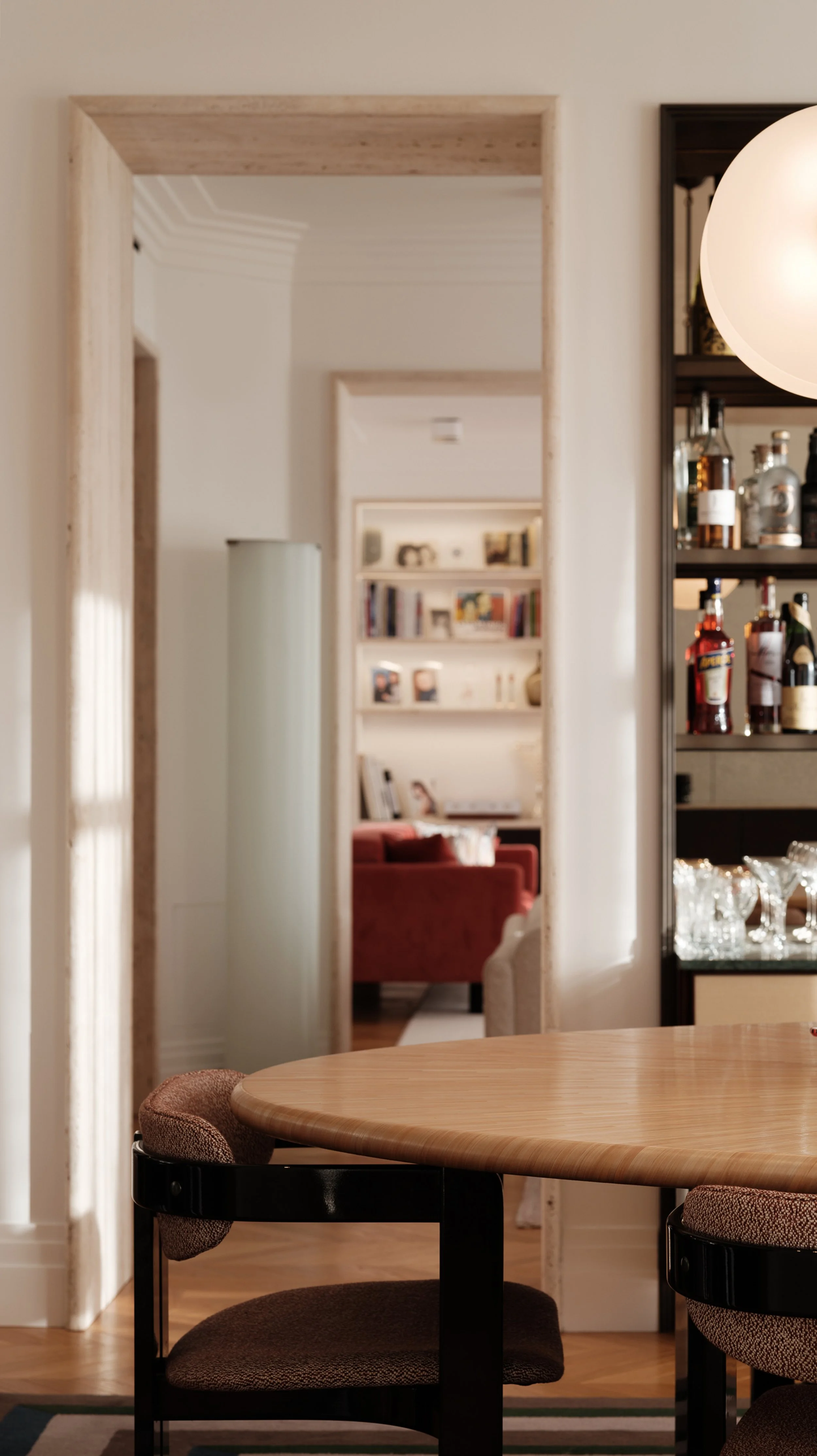 A view through an open doorway shows a dining area with a round wooden table and upholstered chairs in the foreground, a bar with bottles and glassware to the side, and a cozy living room with a red sofa and bookshelves in the background.