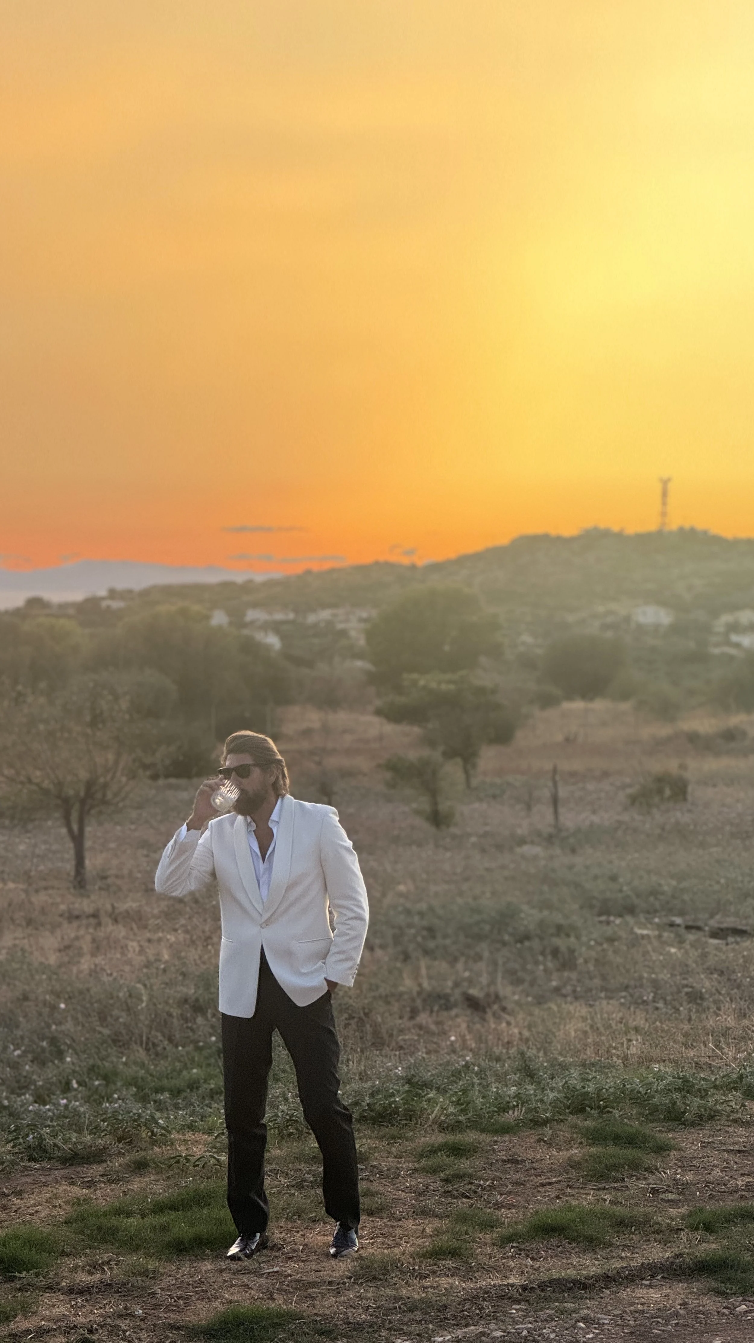 A man in a white blazer and black pants, wearing sunglasses and drinking from a glass, standing outdoors during sunset with a scenic landscape of trees and distant hills.