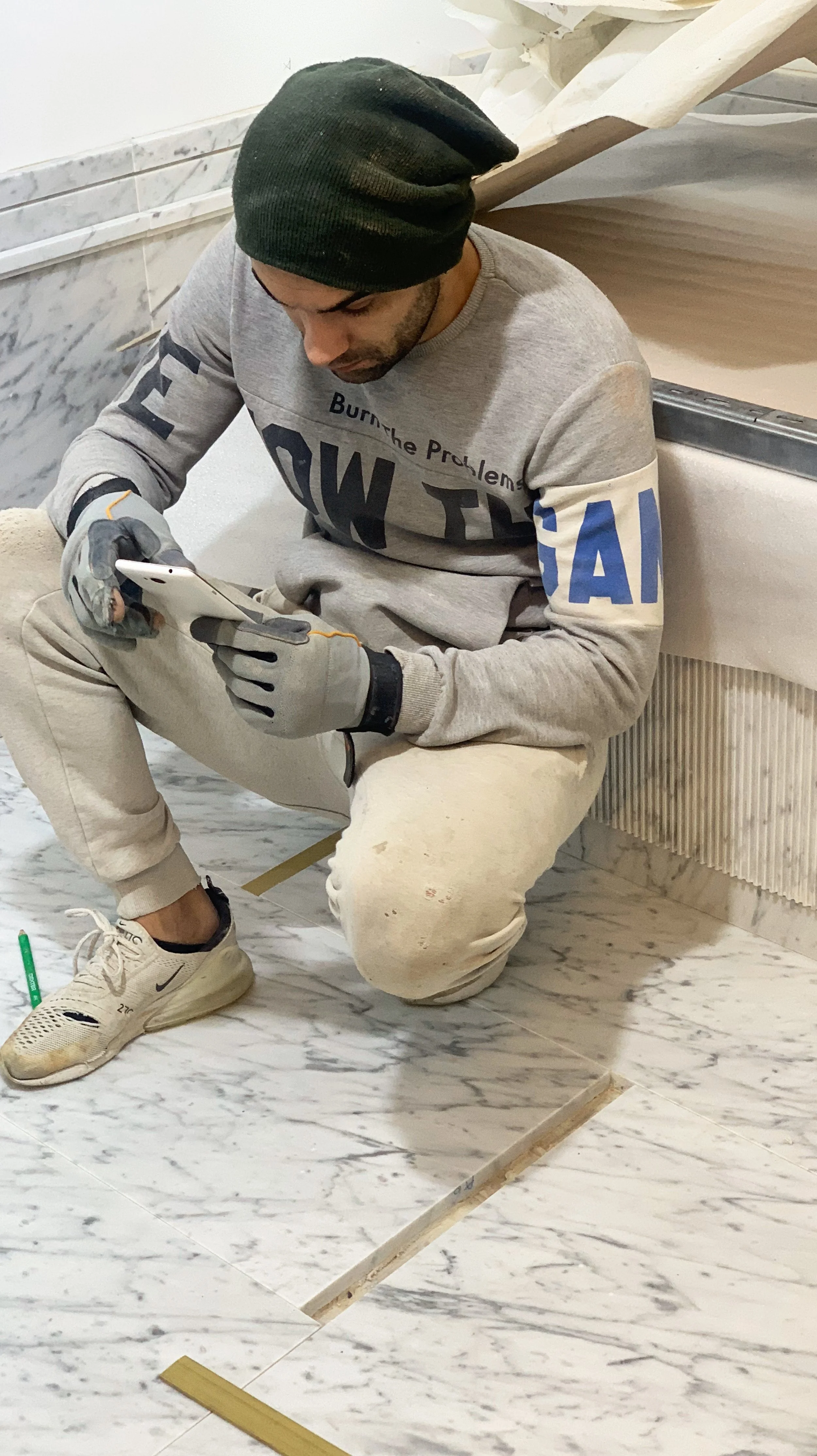 Man in a gray sweatshirt and white sneakers kneeling on marble floor, looking at his phone, with construction tools and materials around him.
