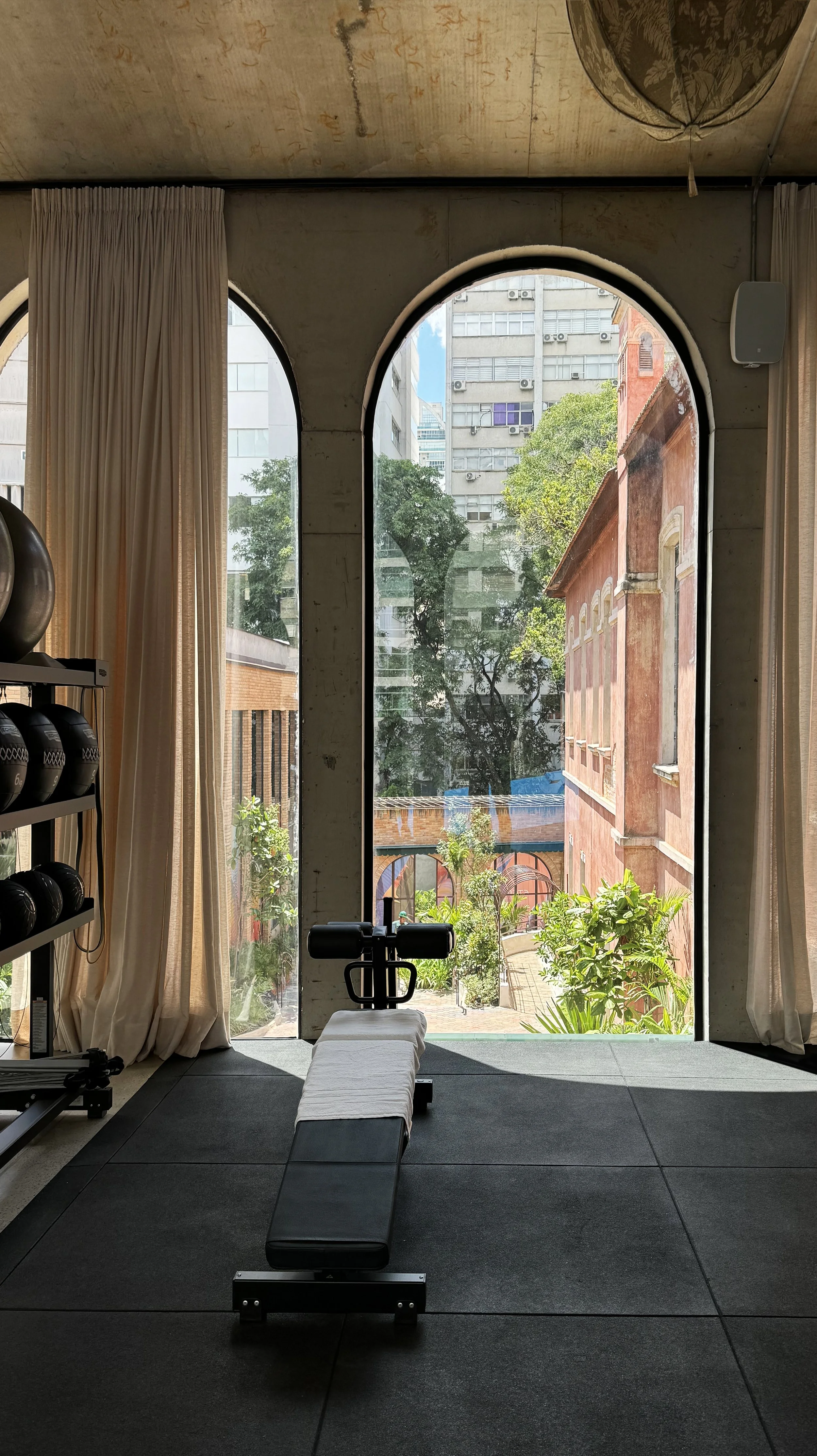 Empty gym with workout equipment, bench, and large arched windows showing city buildings and greenery outside.