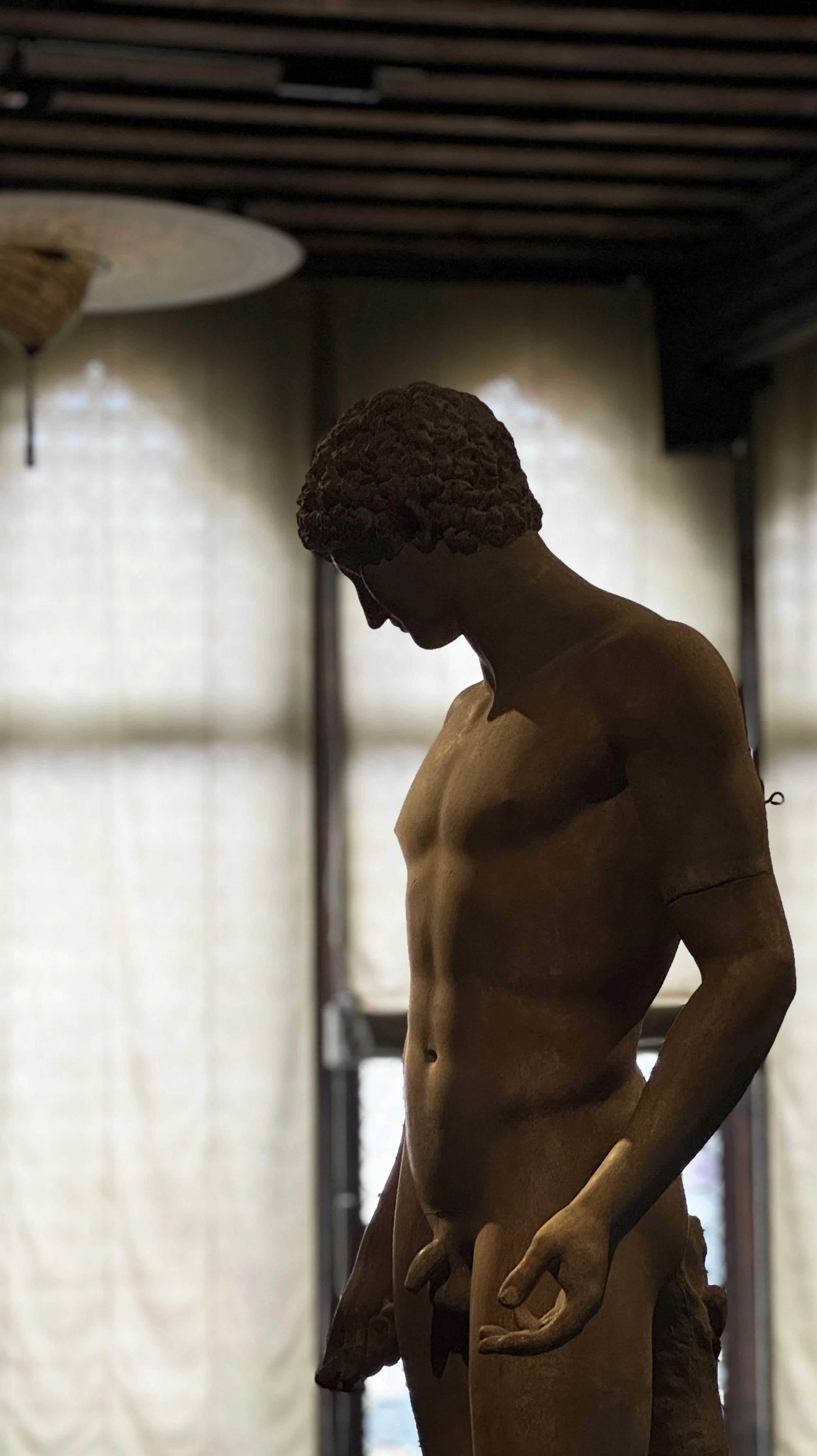 A classical sculpture of a muscular male figure with curly hair, standing indoors with light coming through windows behind it.