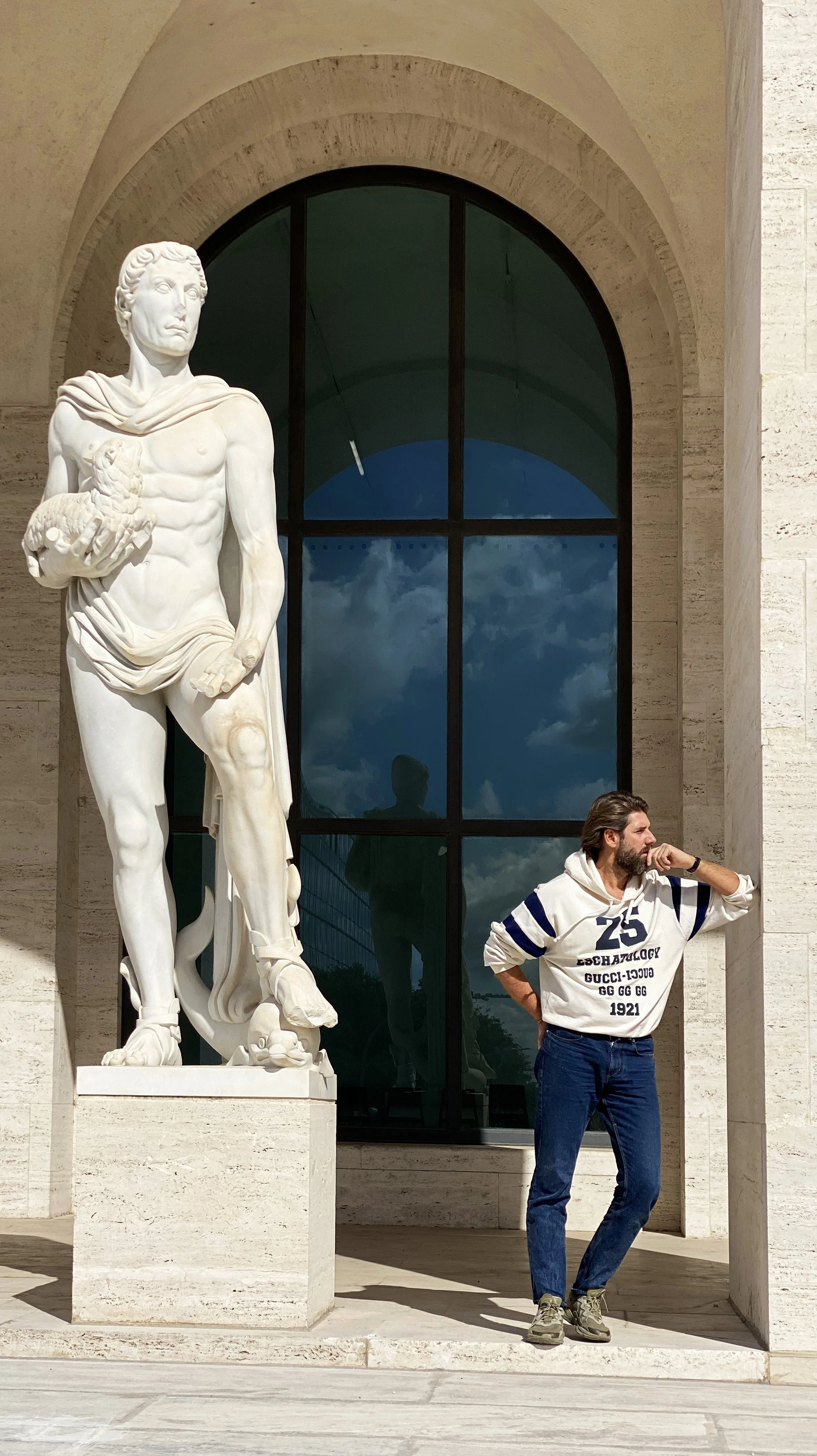 A man standing next to a large classical marble statue of a male figure holding a lion cub, outside a building with a large arched window reflecting a cloudy sky.