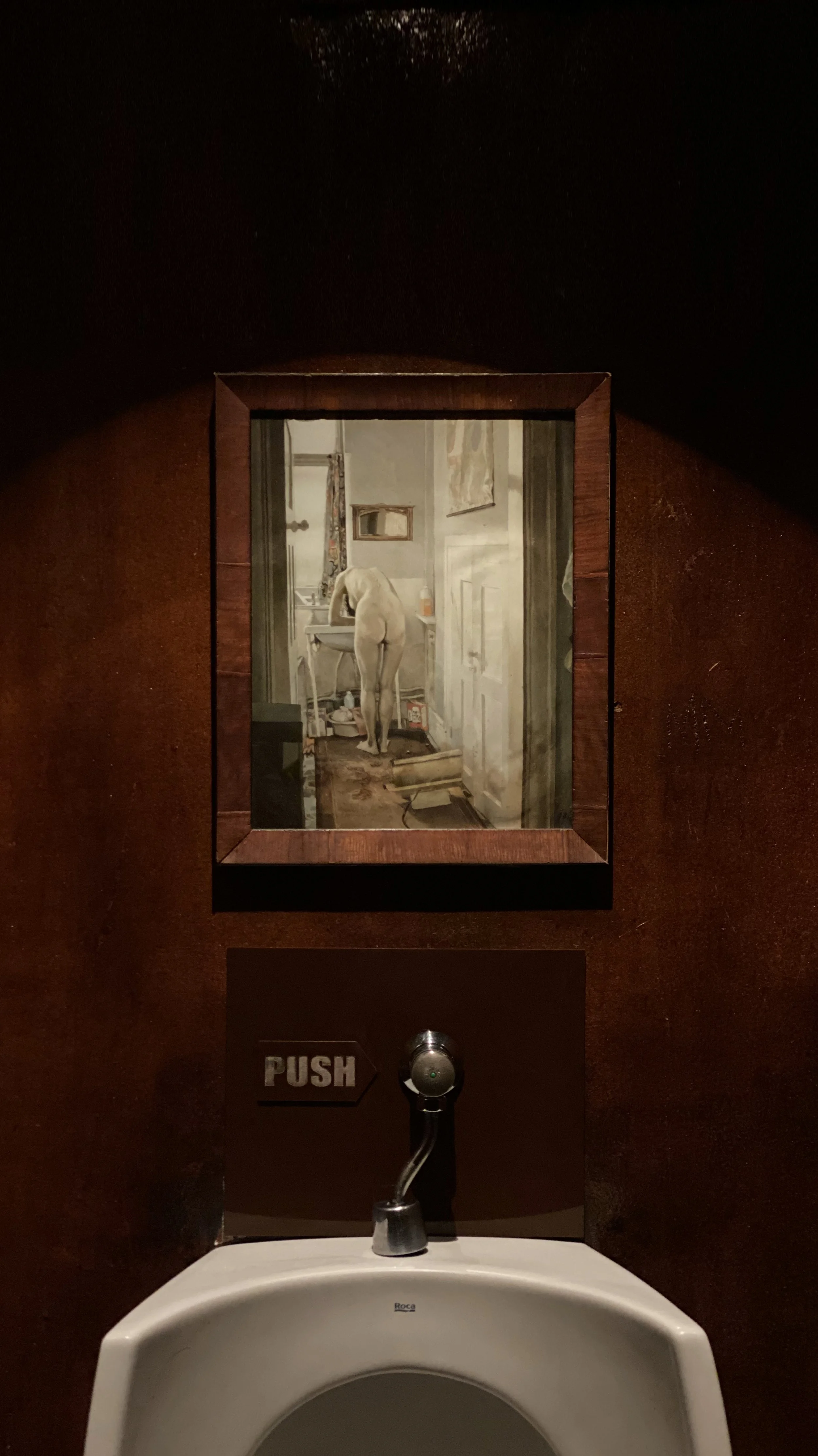 A framed photograph on a bathroom wall depicts a person standing in front of a mirror in a cluttered room, with their back turned and their head bowed down.