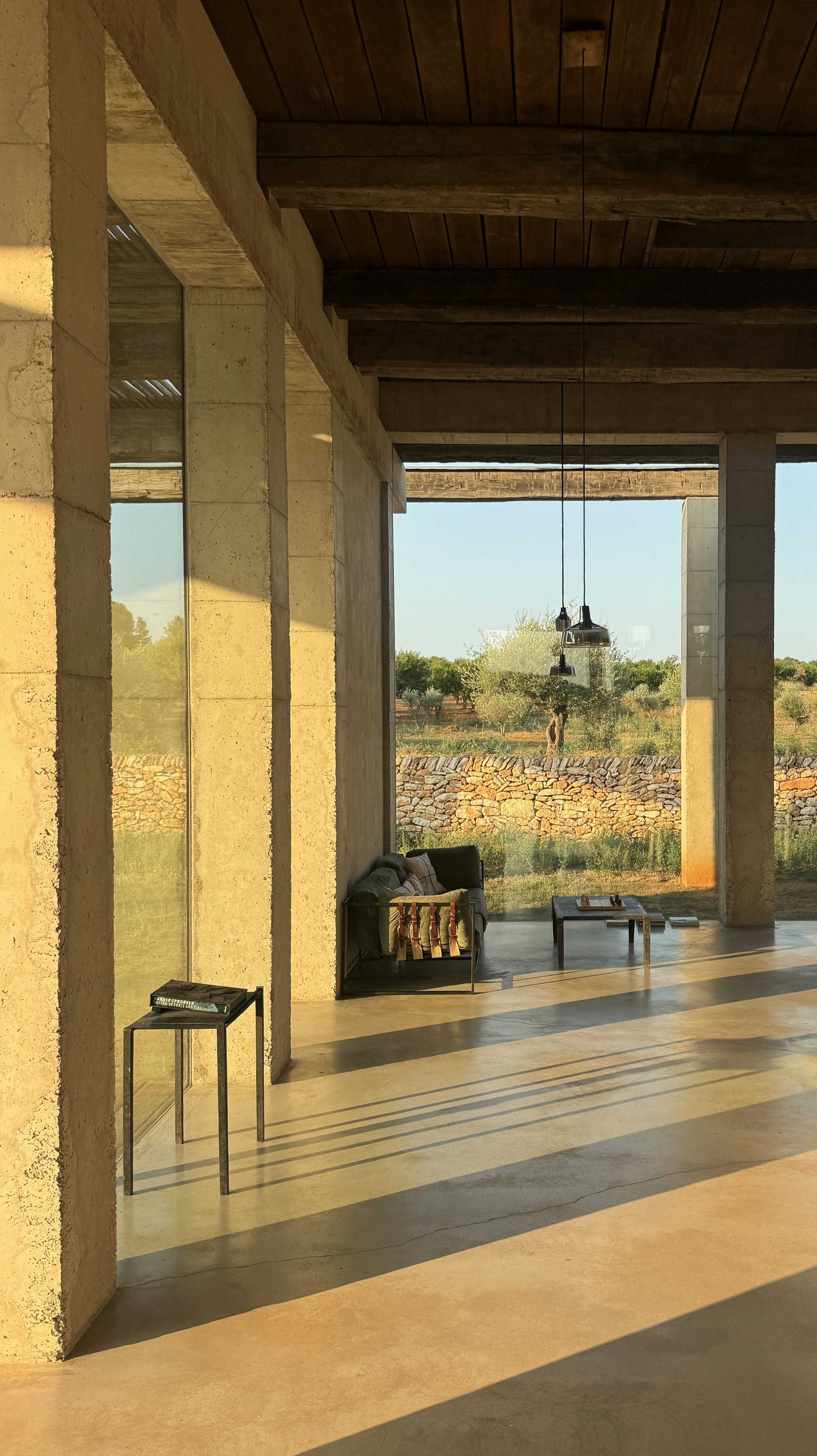 Interior of a modern house with large floor-to-ceiling windows, a concrete floor, and exposed wooden ceiling beams. Sunlight streams inside, casting shadows. There's a small side table with a book, a black sofa with cushions, and a coffee table. Outs