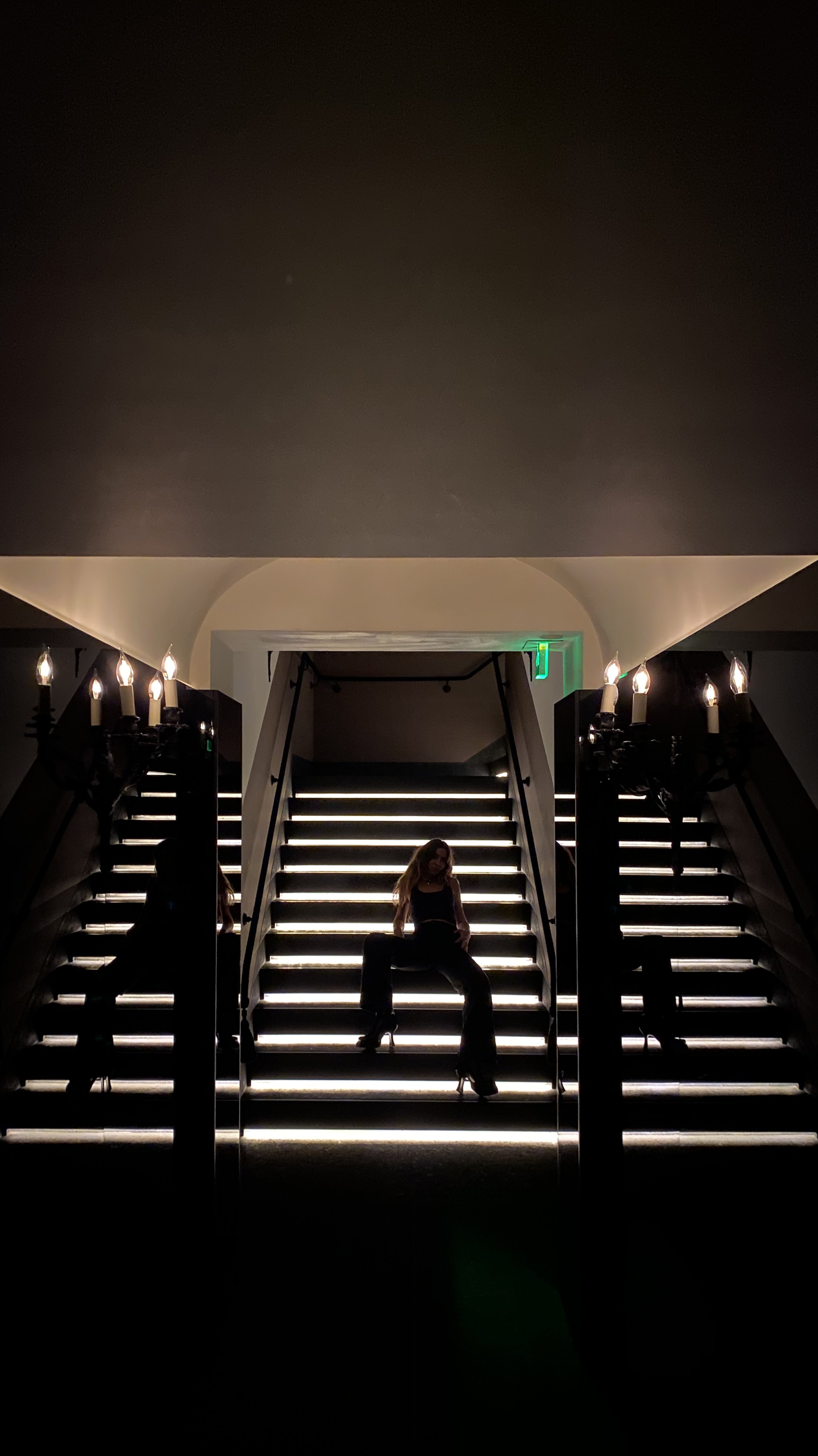 A woman in dark clothing sitting on illuminated stairs with black railings, flanked by wall-mounted candle holders with lit candles, in a dimly lit interior space.