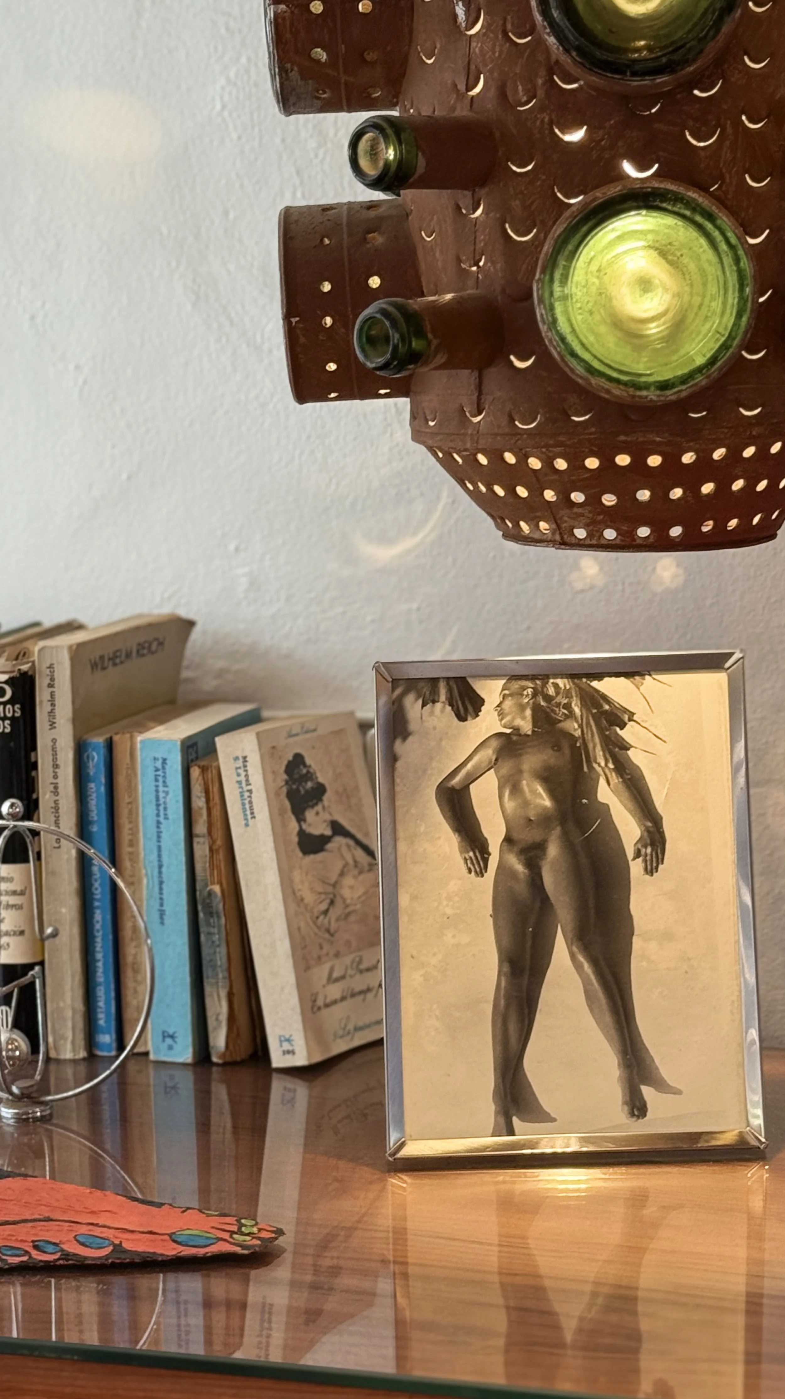 A framed black and white photo of a nude woman with a butterfly on her shoulder, standing with her hands on her hips, is placed on a wooden desk. Behind the frame, there are several books stacked upright, and a decorative pottery piece with small hol