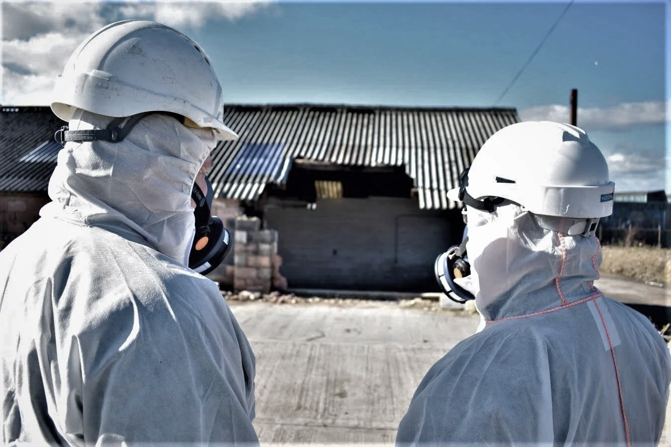 Two workers in white protective suits, helmets, and respirators standing outdoors in front of a damaged building with a corrugated asbestos roof.
