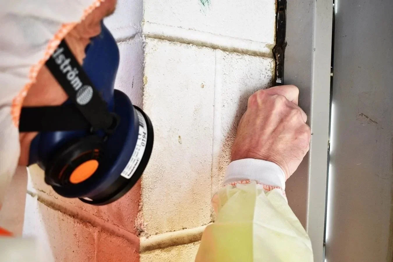 Person performing asbestos testing work on a wall, using a tool; wearing a mask and a white coverall.