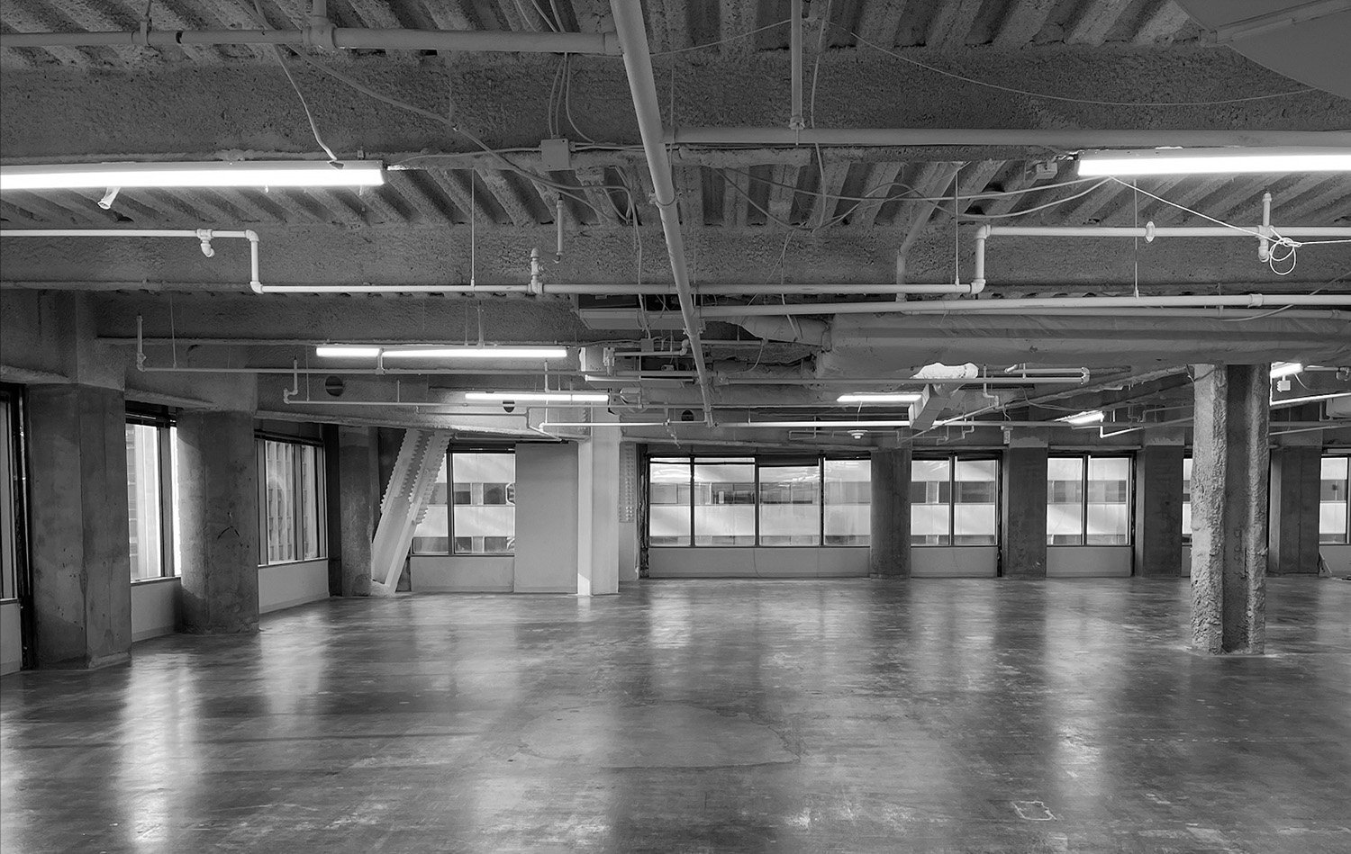 Empty industrial-style room with large windows, concrete columns, polished wooden floor, and exposed ceiling with pipes and fluorescent lights.