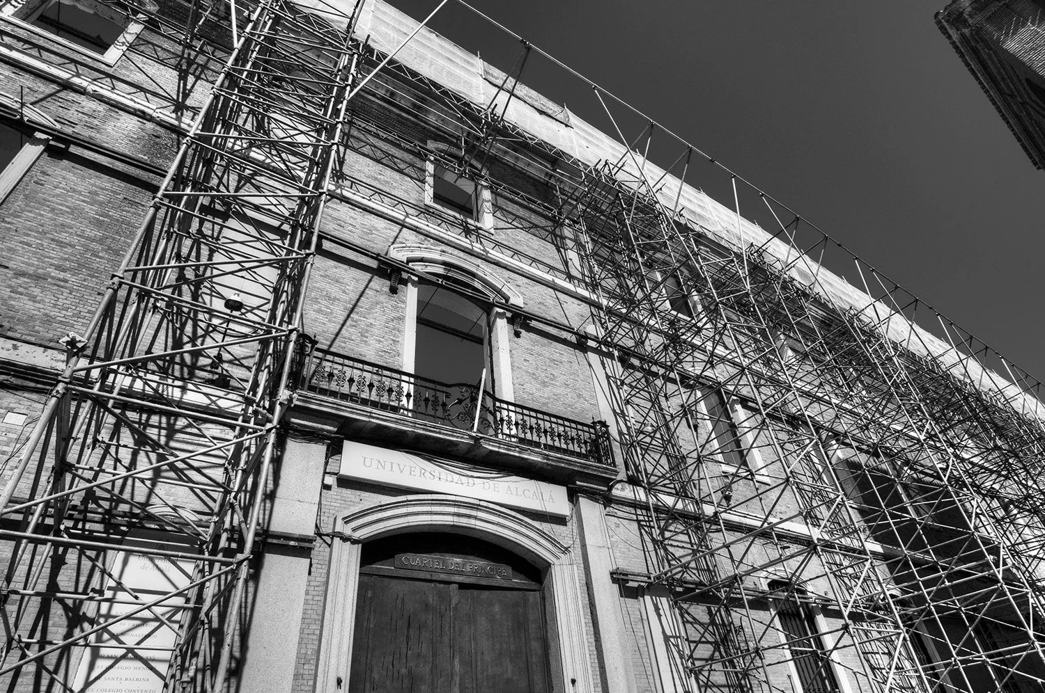 Black and white photo of a historic brick building under construction or renovation, surrounded by scaffolding. The building has arched windows and a sign that reads "UNIVERSIDAD DE ALCALÁ."