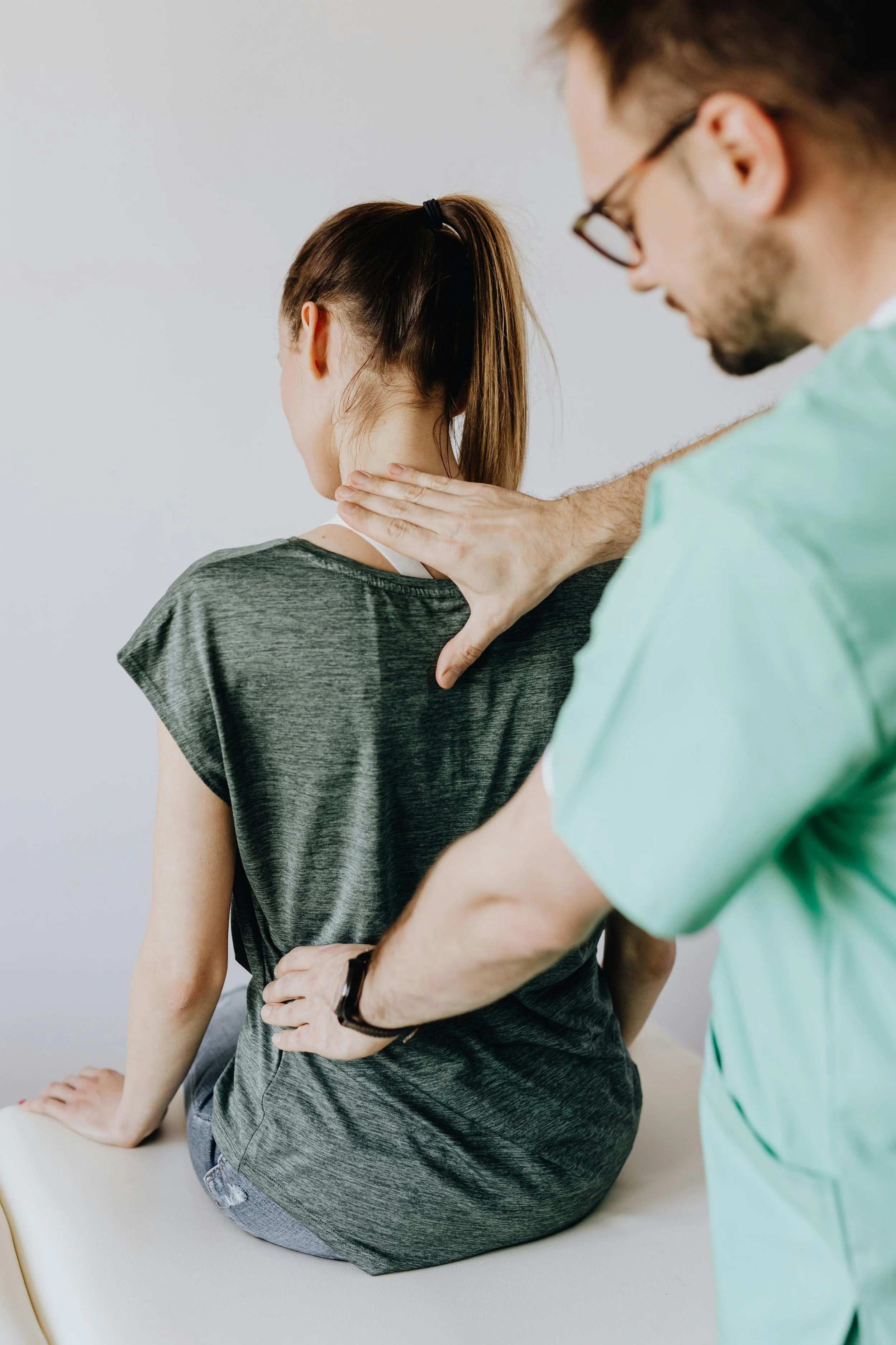 A woman sitting on an examination table experiencing neck pain while a healthcare professional examines her neck and shoulder.