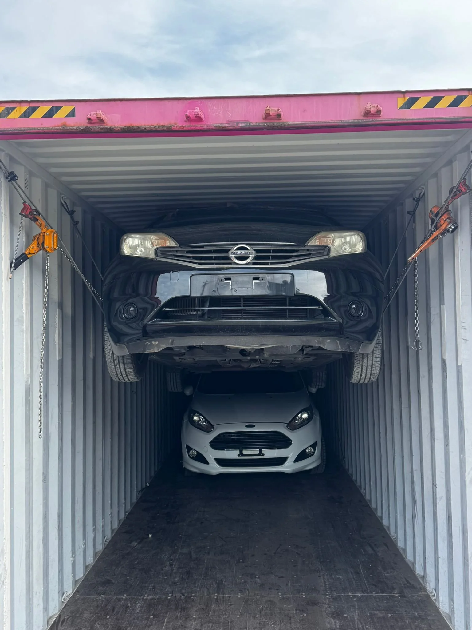 A car transporter trailer with two cars inside: a black Nissan on the top and a white Ford on the bottom.