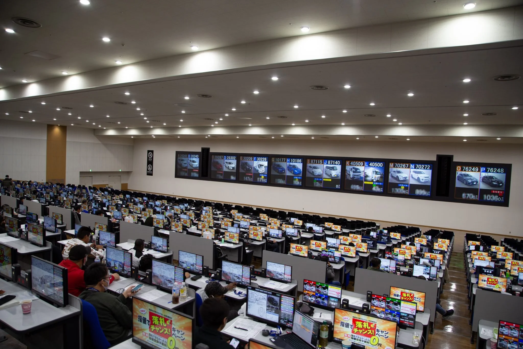 A large room filled with rows of computer desks and monitors, people working at the desks, and a display screen showing different cars and prices.