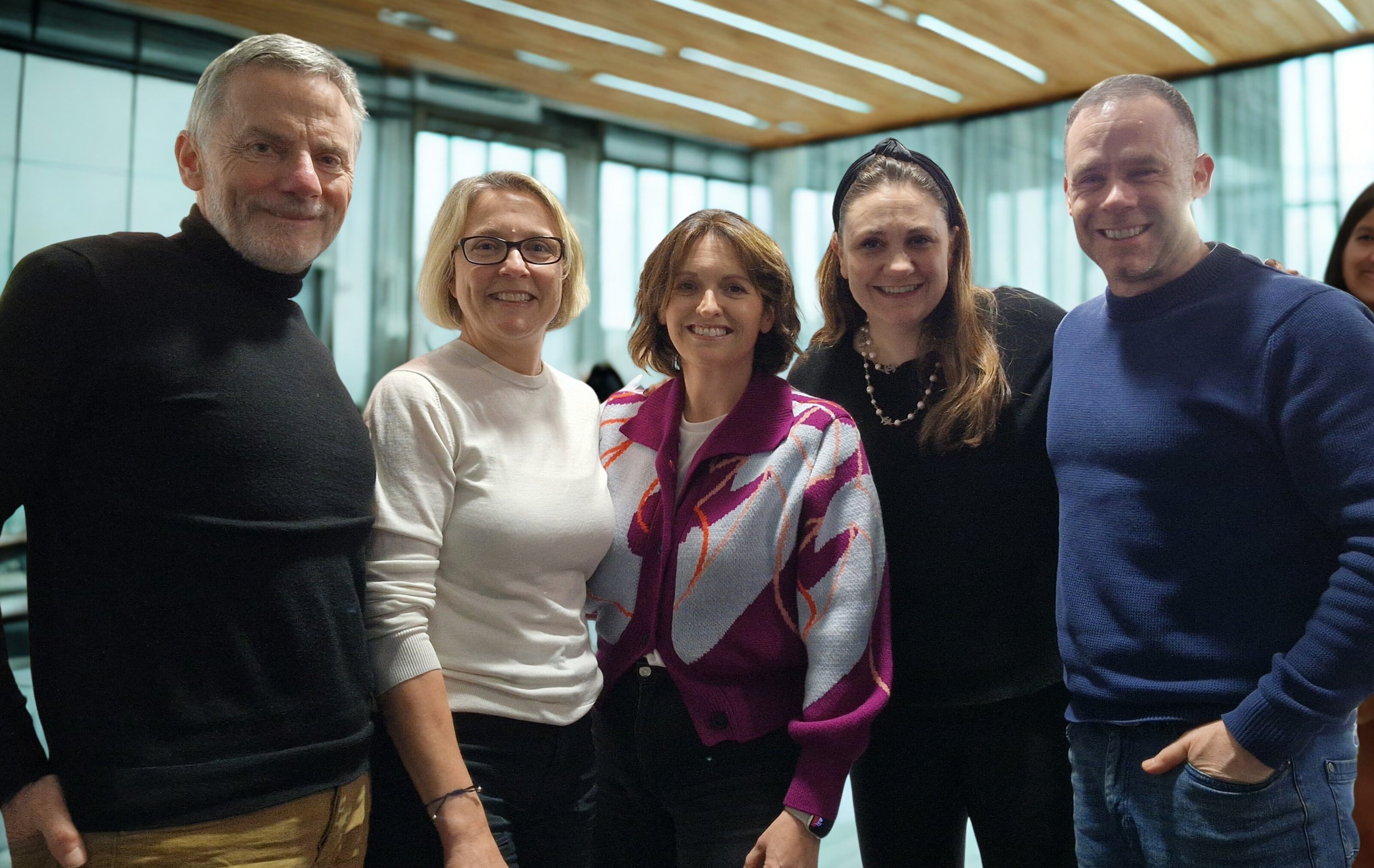 A group of five smiling adults standing indoors in front of large glass windows with a wooden ceiling, posing for a photo.