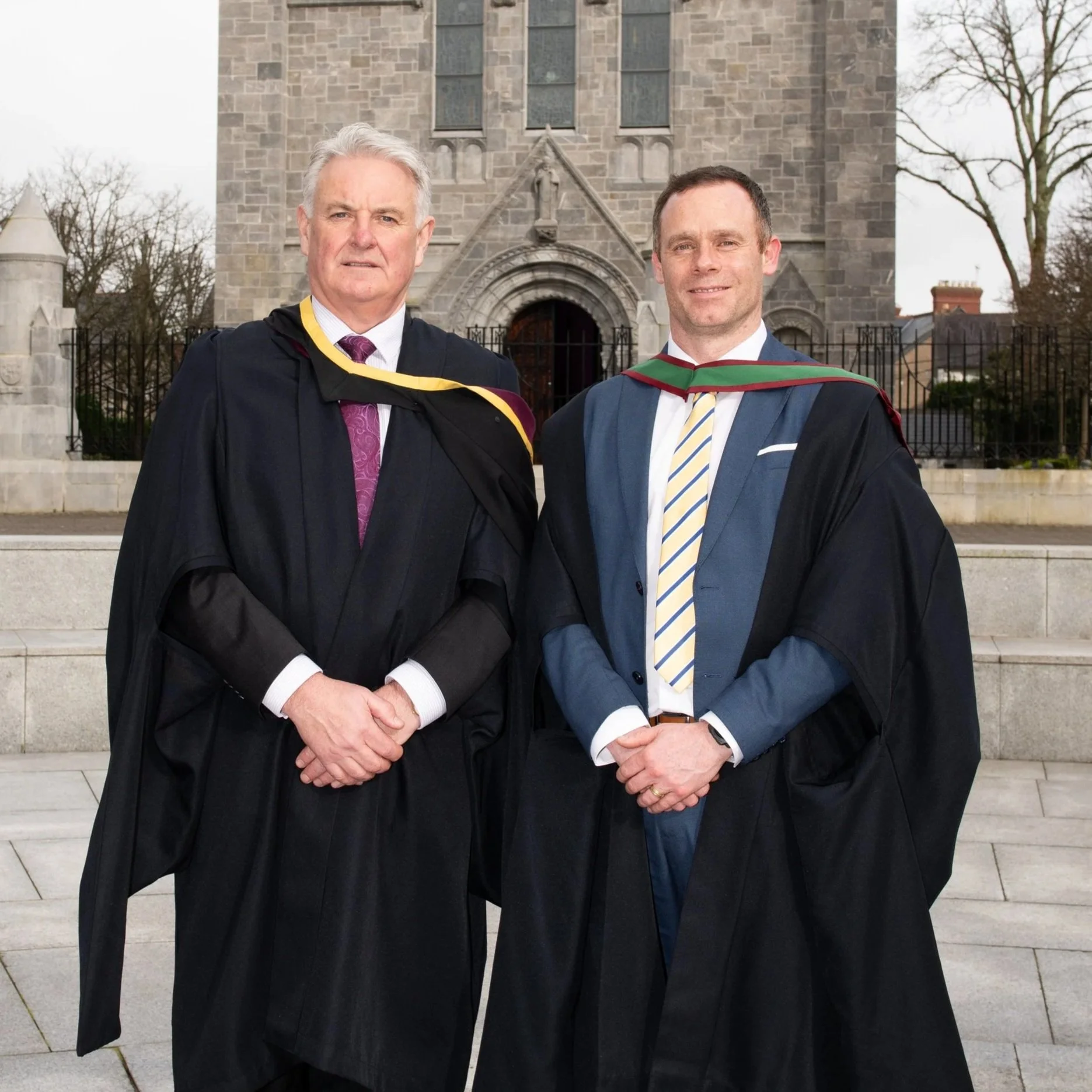 Two men in academic robes standing outside a stone building with a Gothic arch entrance, in front of a metal fence and trees.