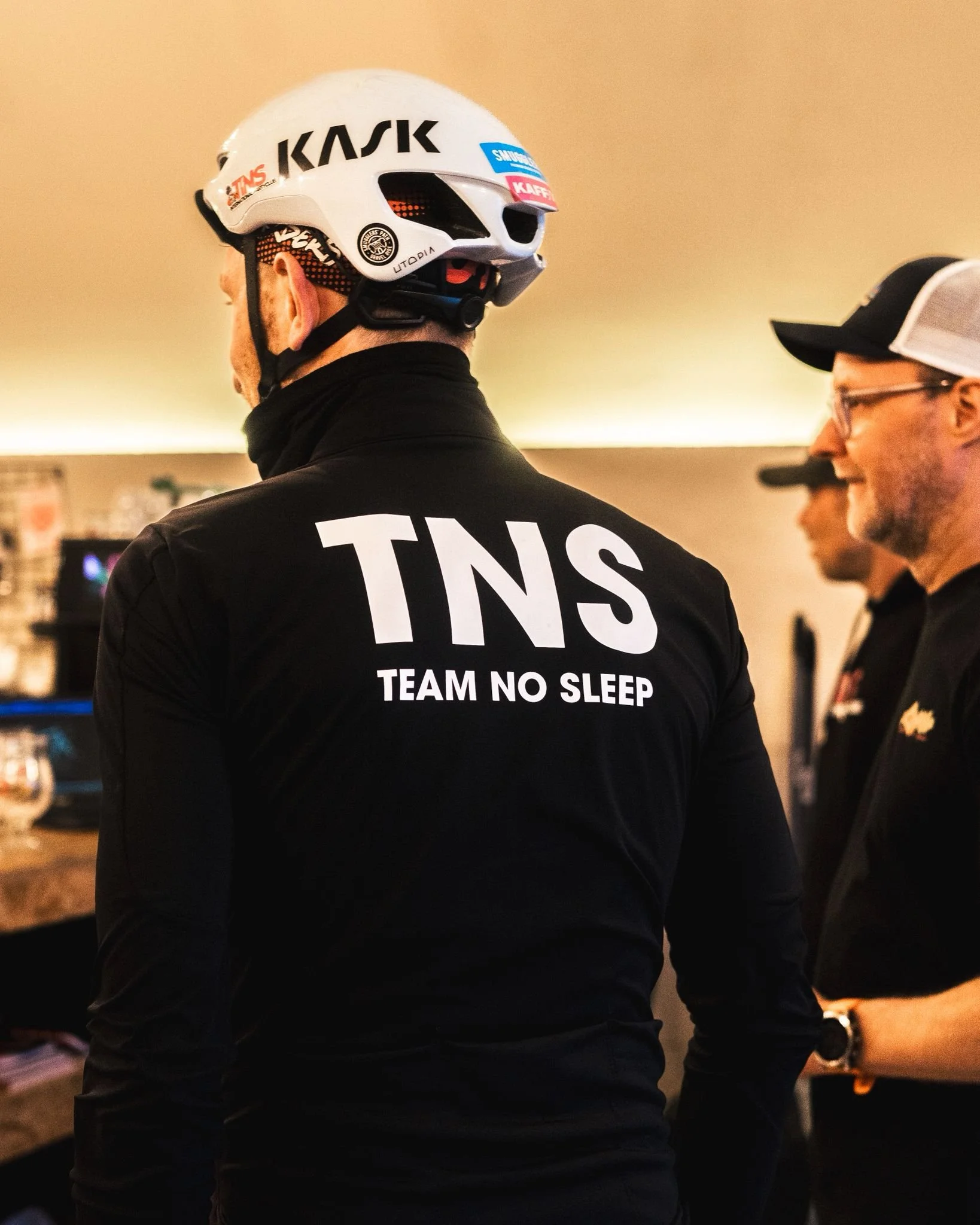 Mannen in een zwart TNS TEAM NO SLEEP-trui met helm die in een binnenomgeving staat, naast een man met een pet en bril.