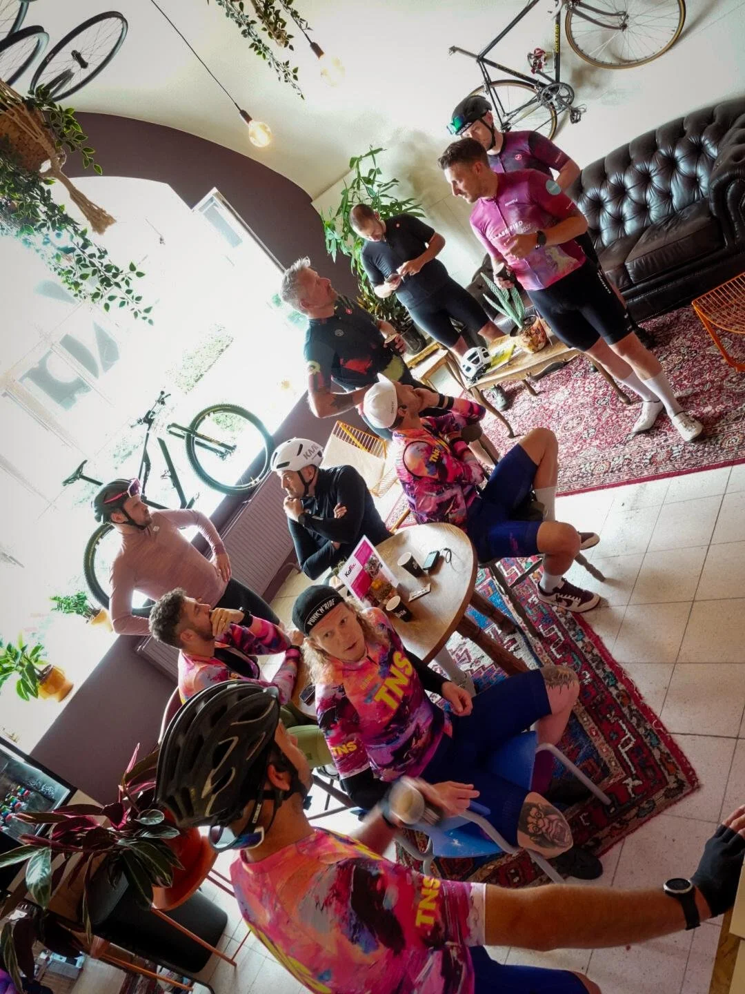 Mensen die zich voorbereiden op een fietsevenement, met enkele in wielrenkleding en helmen, binnen een interieur met fietsen aan de muur, planten en een tapijt.